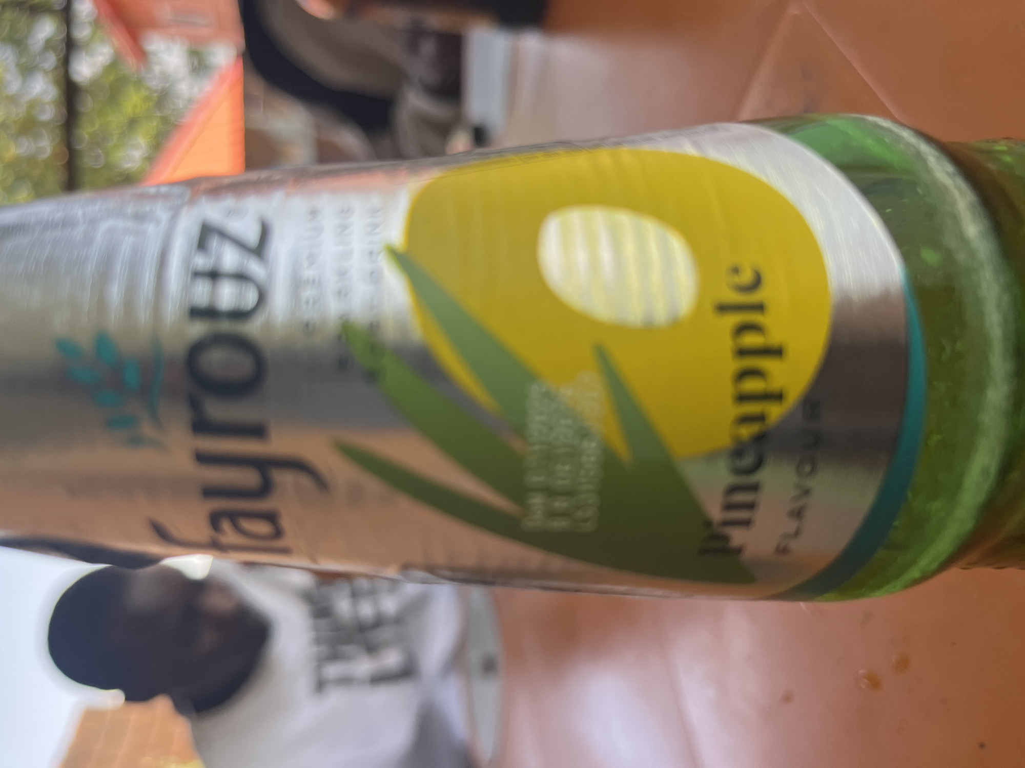 The image shows a close-up, sideways shot of a Fayrouz brand sparkling drink bottle with a pineapple flavor. The bottle is green and the label is predominantly silver and yellow, featuring a stylized pineapple design. Text visible on the label includes "Fayrouz," "PREMIUM SPARKLING DRINK," and "Pineapple FLAVOUR." In the background, out of focus, a person's head and shoulder are visible, wearing a white t-shirt. The setting appears to be outdoors, possibly a casual gathering, given the blurred background elements which suggest trees and an orange surface that could be a table. The lighting is bright, suggesting it's daytime. No specific weather conditions can be determined. The image is grainy and appears to be taken with a phone camera. The overall impression is a candid shot capturing a refreshing beverage in a relaxed environment.