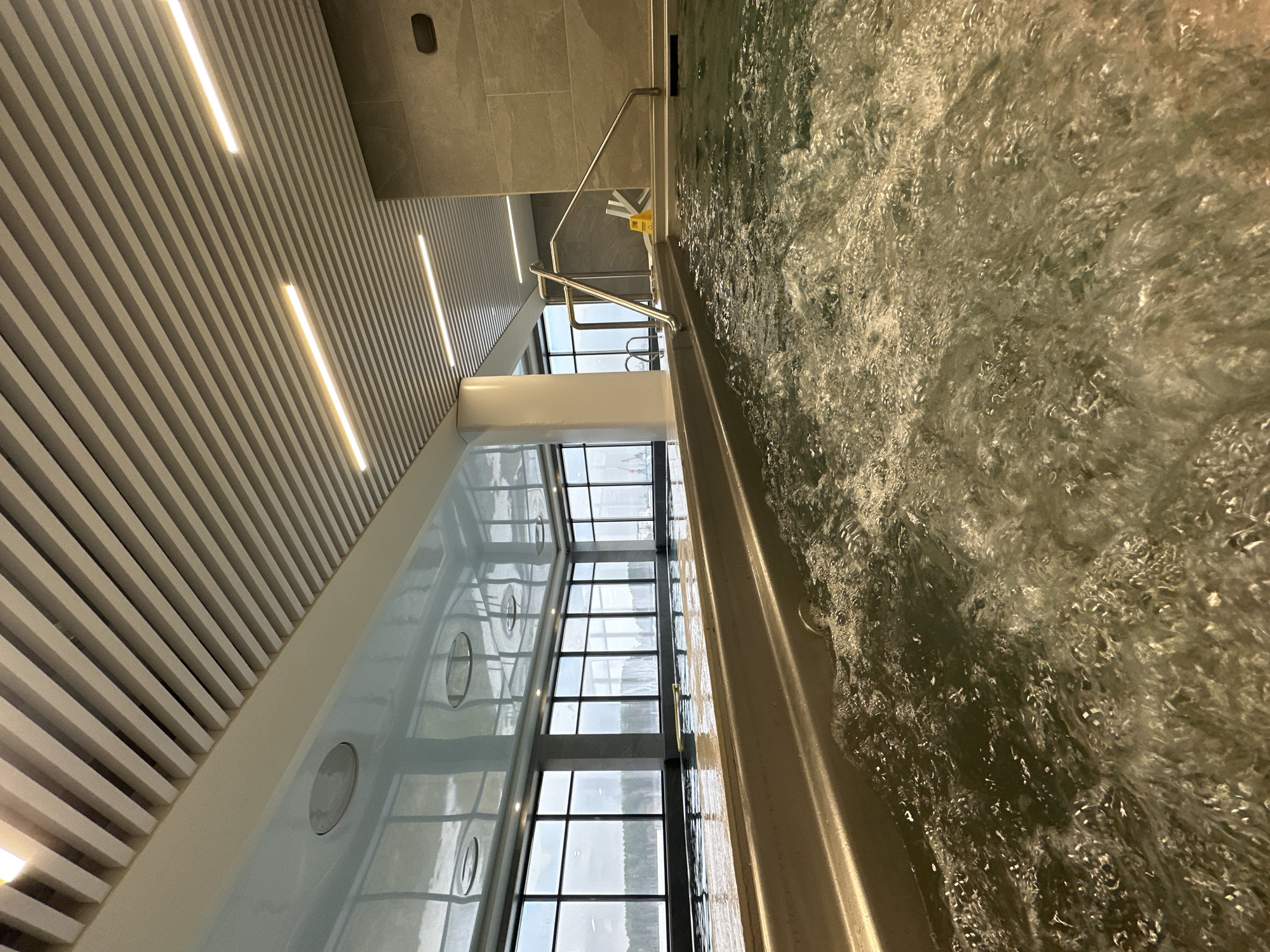 The image captures a perspective from within a hot tub or jacuzzi. The water is bubbly and appears agitated, possibly due to jets. A stainless steel handrail is visible along the edge of the tub. A wall with a large window spans the length of the image, reflecting the cityscape. The architecture indicates a modern or contemporary design. The ceiling is slatted with linear lights embedded within. Due to the reflection in the window, it's impossible to discern the location in Chaikovsky, Russia from visual cues within the image. There are no people visible in the frame. A yellow wet floor sign is located between the wall and the tub.