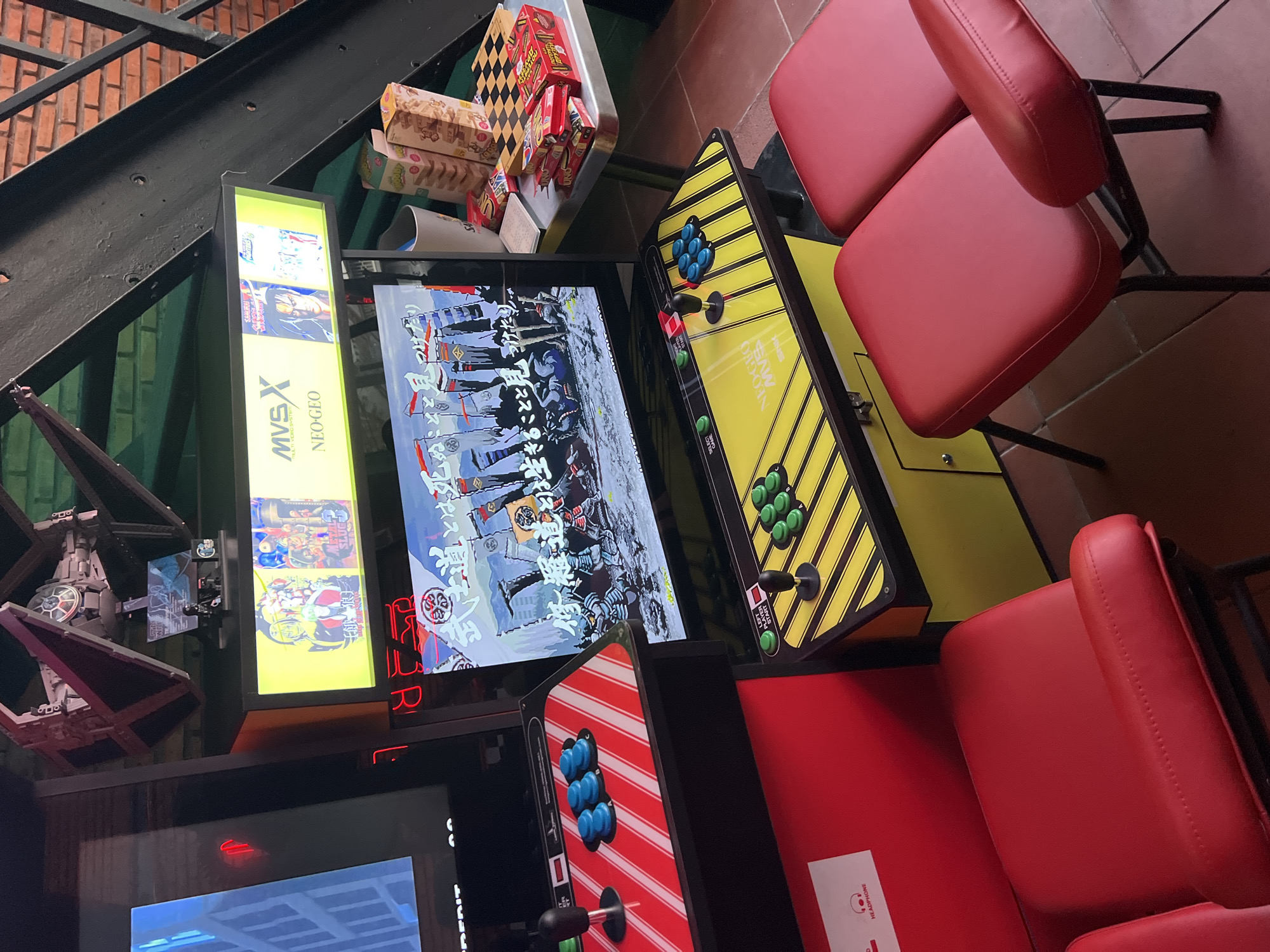 The image depicts a corner of an arcade with several arcade game cabinets. The location appears to be in Bac Nha Trang Ward, Vietnam based on the OCR data.
* **Arcade Cabinets:** There are several arcade cabinets, including one with a yellow panel and one with a red panel. The yellow cabinet has diagonal black stripes on the panel and green buttons. The red cabinet has vertical white stripes on the panel and blue buttons. One cabinet has a display screen showing a samurai-themed video game. The cabinet on the right has two red padded bar stools. Another cabinet has an illuminated sign that says "MVSX NEO-GEO" on the side.
* **Other Objects:** A model of a Star Wars TIE fighter is visible on top of one of the cabinets. There are also boxes of snacks on a shelf above the cabinets.
* **Environment:** The location is indoors, possibly a bar or restaurant with an arcade corner. The background includes a brick wall and stairs.
* **People/Actions:** There are no people visible in the shot, and no actions are depicted.