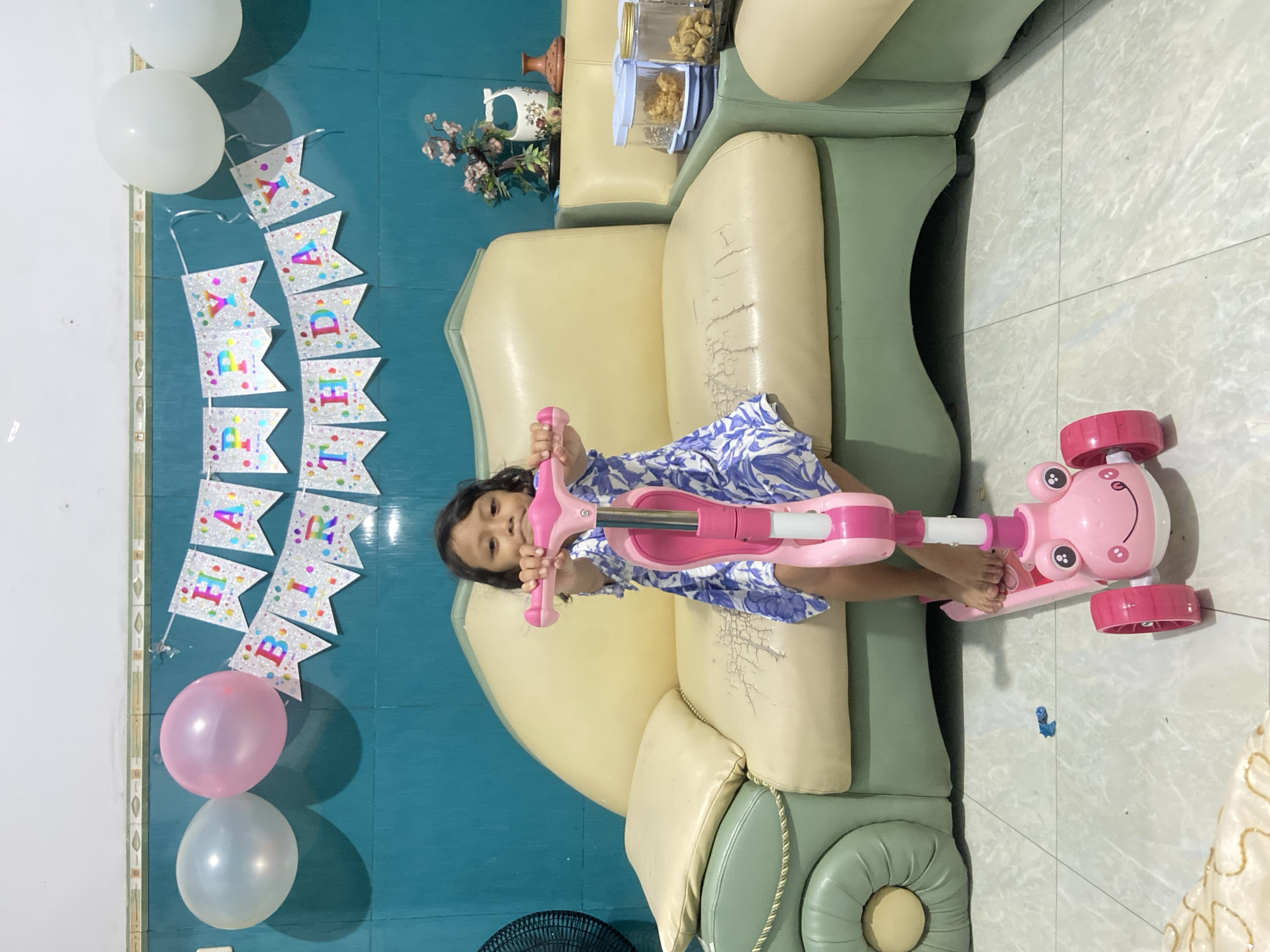 A young girl in a blue and white dress is sitting on a light-colored, slightly worn sofa. She is holding a pink scooter. Behind her, a "HAPPY BIRTHDAY" banner and balloons are affixed to a blue wall. A corner of a fan is visible in the lower left, and a portion of a white tablecloth is visible in the lower right. A multi-tiered sofa with a vase of flowers is placed near a wall. The location is likely a residence in Gelam, Indonesia.