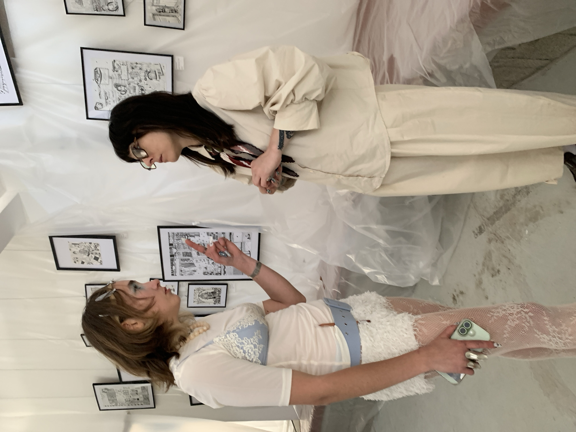 Two women are standing in an art gallery, possibly in Tel-Aviv, Israel, considering artwork on the walls. The gallery space is decorated with white plastic sheeting. One woman is wearing a light beige suit. The other woman is wearing a white ensemble with lace detailing and a light blue belt. She is holding a smartphone. The walls are lined with framed black and white drawings.