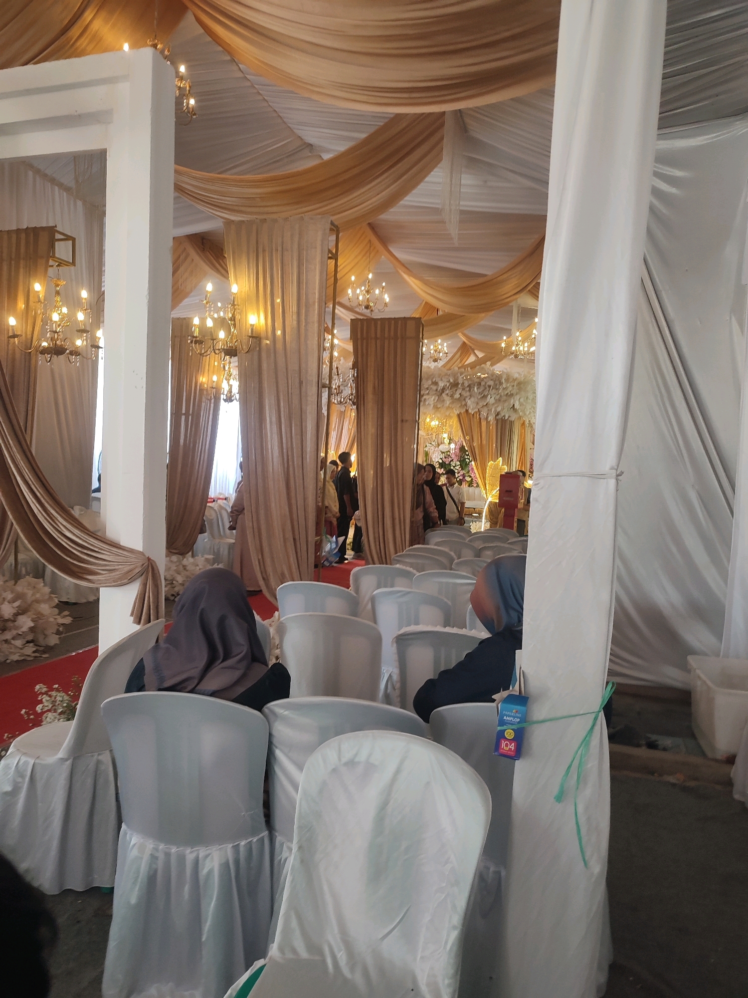 The image depicts an indoor event space, likely a venue for a wedding or similar celebration, within Kecamatan Haurgeulis, Indonesia. The setting is decorated with flowing gold and white drapes suspended from the ceiling, creating an elegant ambiance. Several ornate chandeliers with multiple bulbs are visible, illuminating the area. Rows of white-covered chairs are arranged, suggesting seating for guests. In the foreground, two individuals are seated, both wearing headscarves. One person is visible from behind, wearing a dark headscarf and dark clothing. The other person, also facing away, wears a light blue and grey headscarf and a dark top. Further back, a group of people can be seen standing, some partially obscured by drapes and columns. One individual in this group is wearing a light-colored dress, and another is dressed in dark clothing. The overall impression is of a formal gathering in progress or about to begin. A blue tag with some text and numbers, possibly a seating assignment or identifier, is tied to the back of one of the chairs with green string. The floor is covered with a red carpet in the central aisle area. White floral arrangements are visible on the floor near the chairs and in the background.