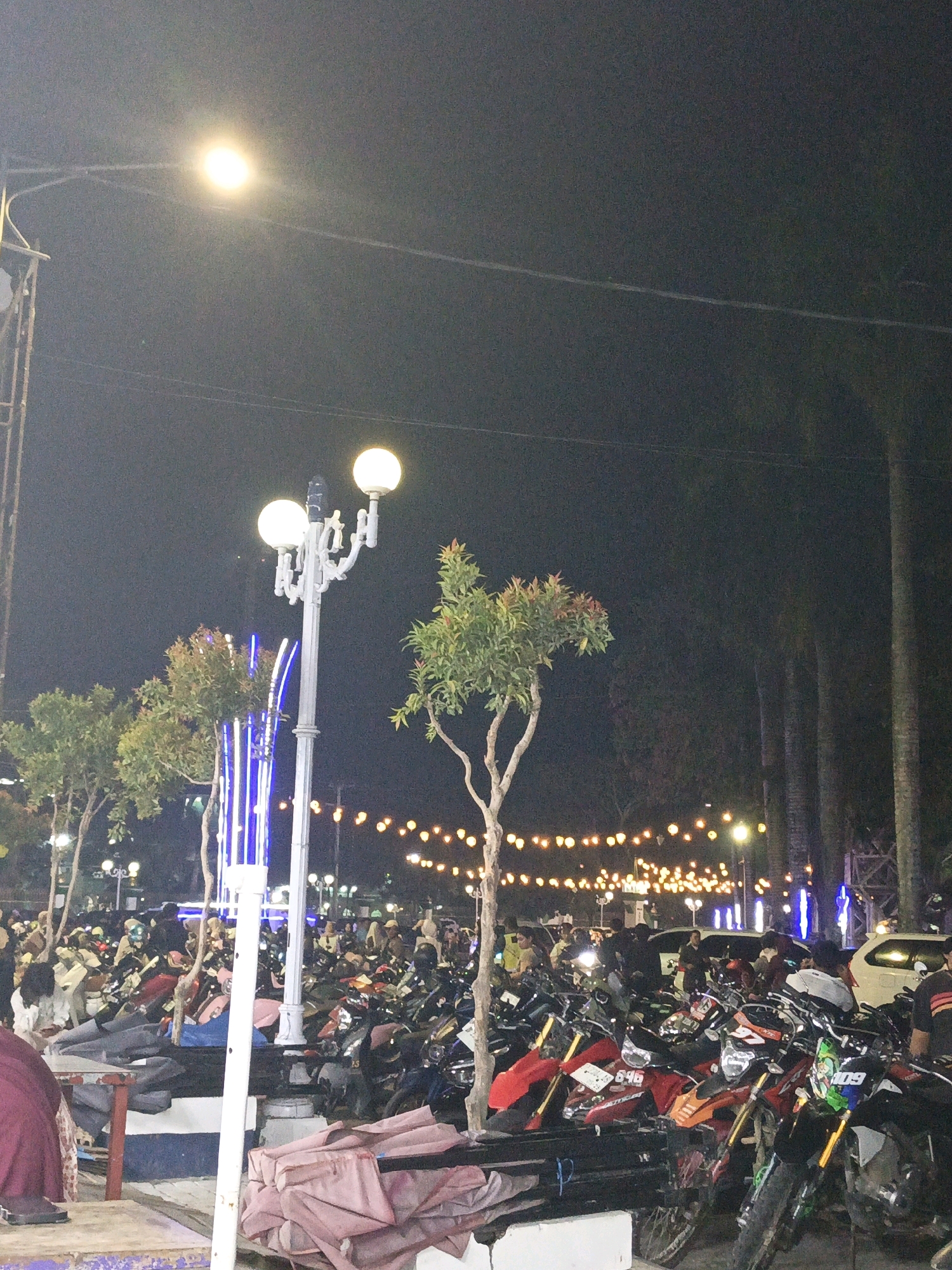 The image depicts a busy night scene at a public parking area, likely in the city of Pangkajene, Indonesia. The foreground features a portion of a table and some stacked pink fabric items. Dominating the mid-ground is a large collection of parked motorcycles, with their headlights and various colors visible under artificial lighting. Several people are dispersed throughout the parking area, some appearing to be retrieving or parking their motorcycles, while others are standing or sitting.
Behind the motorcycles, a decorative lamppost with multiple spherical lights stands prominently. Further in the background, strings of warm-toned fairy lights are strung across, illuminating the gathering and creating a festive atmosphere. Several trees with green foliage are scattered throughout the scene, their shapes outlined by the lights. Tall, slender trees, possibly palm trees, are visible in the far background. The sky is dark, indicating it is nighttime. The overall impression is of a lively, crowded evening event or gathering at an outdoor parking space.