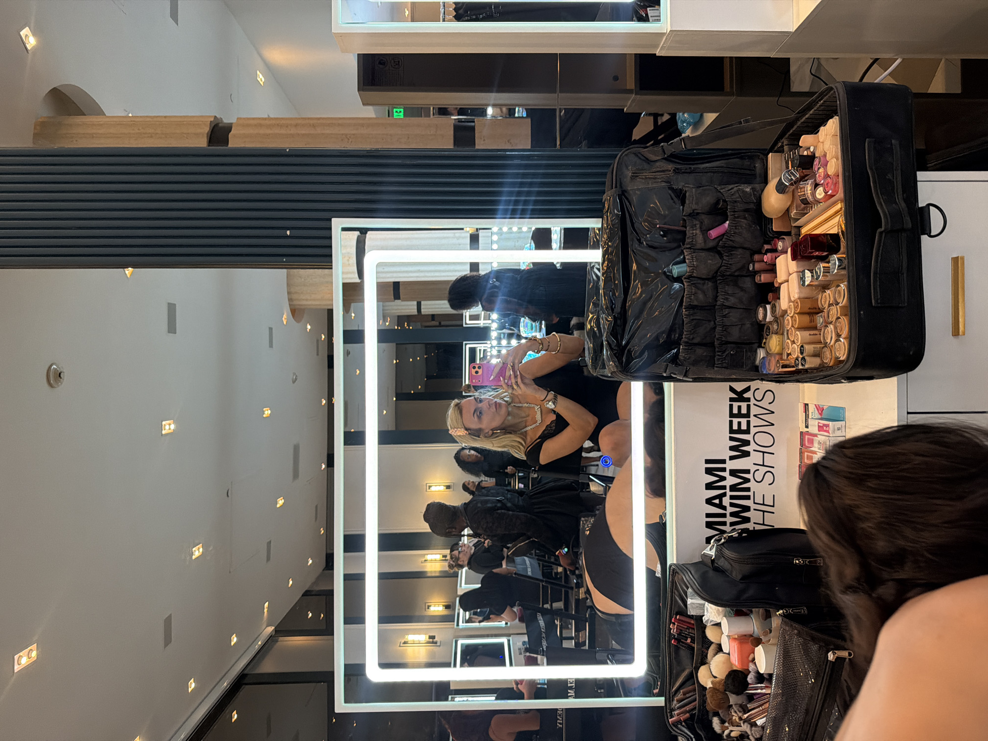 The image depicts a scene in what appears to be a backstage area during Miami Swim Week in Miami Beach, Florida, United States of America. A makeup artist's station with a large illuminated mirror is visible, reflecting a line of individuals, including models and makeup artists, presumably preparing for a show. One model, wearing a black dress and jewelry, is taking a picture of herself in the mirror with a cell phone. The surrounding environment suggests a high-end or professional setting with interior lighting and contemporary design. A makeup case filled with cosmetics is also visible. The text "MIAMI SWIM WEEK THE SHOWS" is printed on a sign near the mirror.