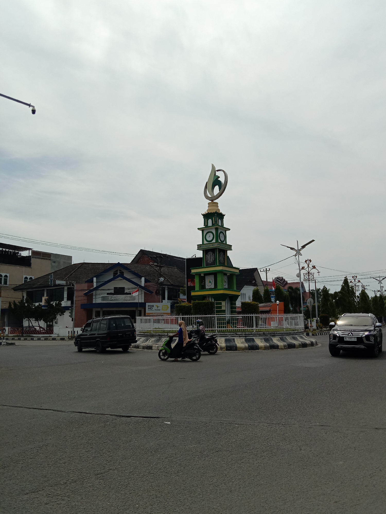 This media file captures a daytime scene at a bustling roundabout in Lhokseumawe, Indonesia, under an overcast sky.
The central feature of the roundabout is a multi-tiered monument, primarily colored green and yellow, which resembles a clock tower. Four clock faces are visible on its upper section. Capping the monument is a distinctive, abstract sculpture with green and gold elements. The base of the monument is encircled by a white metal fence.
Vehicles are present on the road around the roundabout. On the right, a dark-colored Mitsubishi Pajero Sport SUV is seen, with its license plate reading "BL 1109 KI." On the left, a dark van is driving. A person wearing a blue top and hijab is riding a green scooter behind the van, and another dark scooter is partially visible.
In the background, several buildings line the street. Prominently featured is a building with a blue roof and a distinctive facade, identified as "KANTOR POS" (Post Office) by text above its entrance. A sign to its right displays "POSIND" and "POSPAY." Other buildings with varying architectural styles and tiled roofs are visible further back. Street lights and power lines crisscross the upper part of the image, and some trees line the road. The road surface appears to be asphalt.