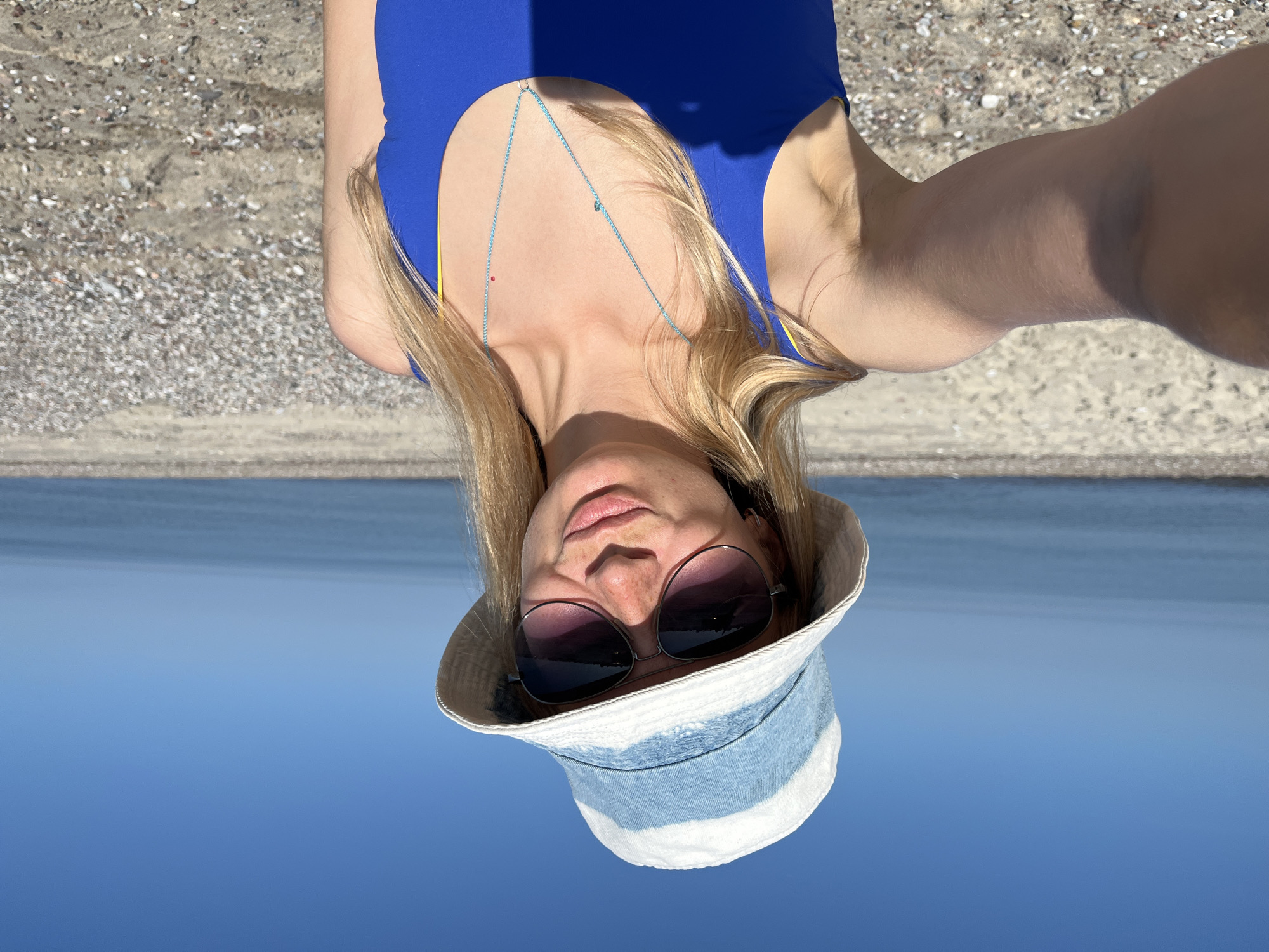 A young woman with blonde hair, wearing sunglasses, a bucket hat with blue and white stripes, and a blue swimsuit, poses for a selfie on a beach. The background shows the blue sky and the sea. The beach appears to be pebbly.