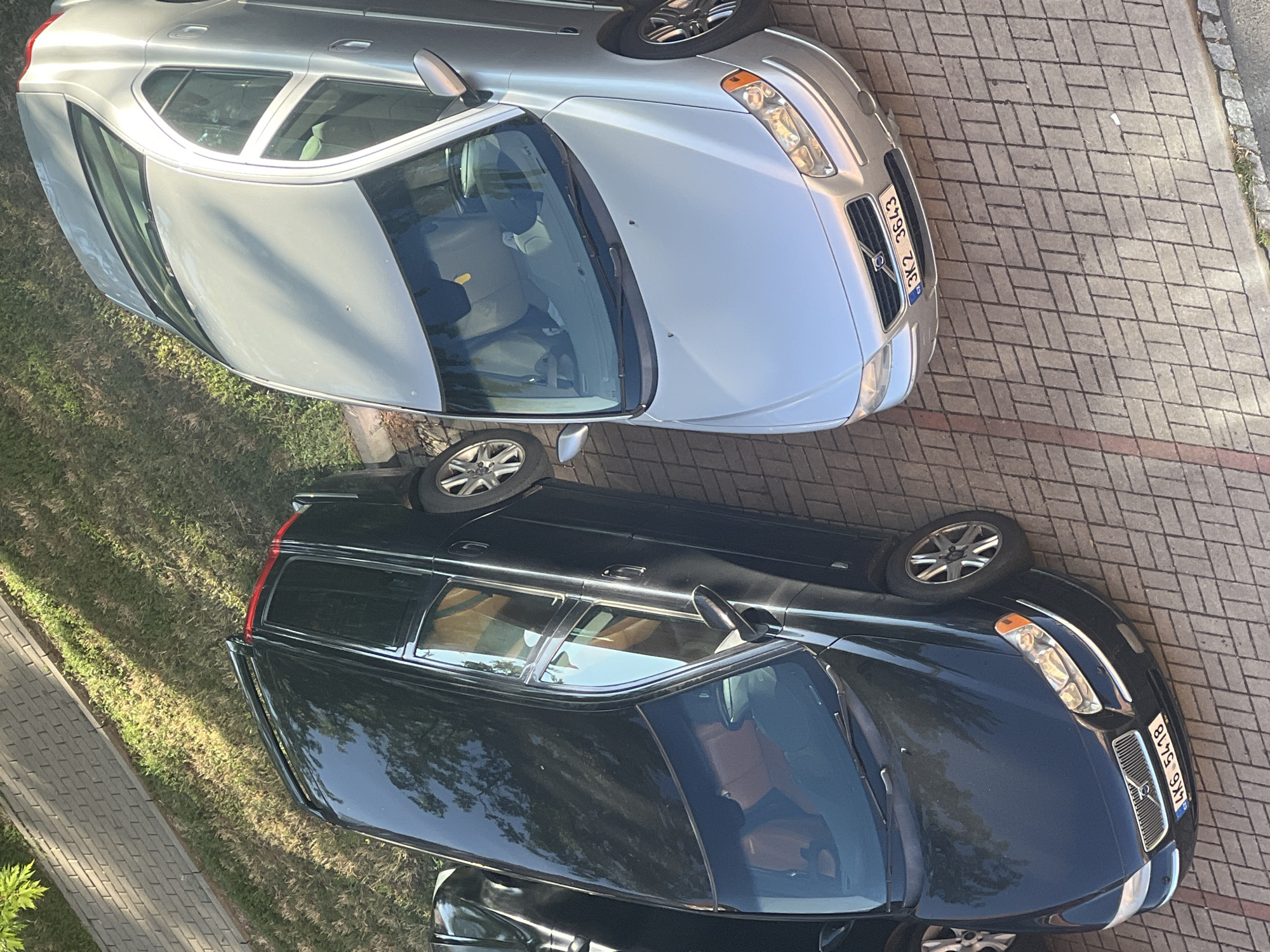 The image shows two Volvo cars parked on a paved surface in Cheb, Czechia. The car on the left is a black station wagon, and the car on the right is a silver sedan. Both cars are angled and viewed from above. The black station wagon has a license plate that reads "4K6 5418". The silver sedan has a license plate that reads "3K2 3643". A strip of green grass borders the paved area on the left side of the image, and some trees are reflected in the windows of the cars. The lighting suggests it is daytime.