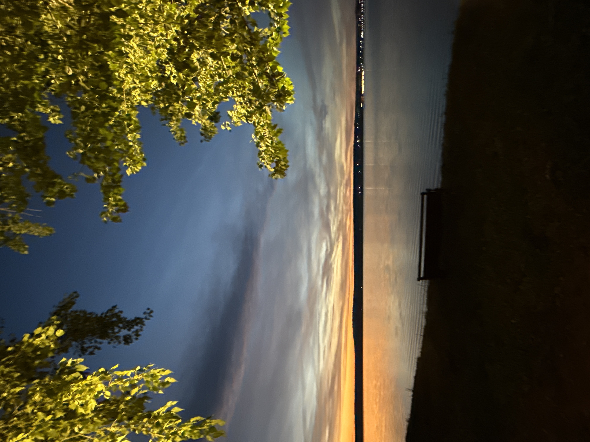 The image depicts a serene natural landscape at dusk, likely in Votkinsk, Russia, given the context. In the foreground, a dark silhouette of a grassy embankment leads down to a body of water. On the embankment, a lone, dark-colored bench faces the water. Overhead, the branches of a tree, illuminated from below, create a frame for the scene, with bright yellow-green leaves contrasting against the darkening blue sky. The sky itself transitions from a vibrant orange and yellow near the horizon, indicative of sunset, to a deep blue overhead, with streaks of wispy clouds. The water's surface reflects the colors of the sky, creating a shimmering effect. Across the water, a distant, dark silhouette of a shoreline is visible, dotted with faint lights, suggesting the presence of buildings or a town. The overall atmosphere is peaceful and contemplative, capturing the transition from day to night in a scenic locale.