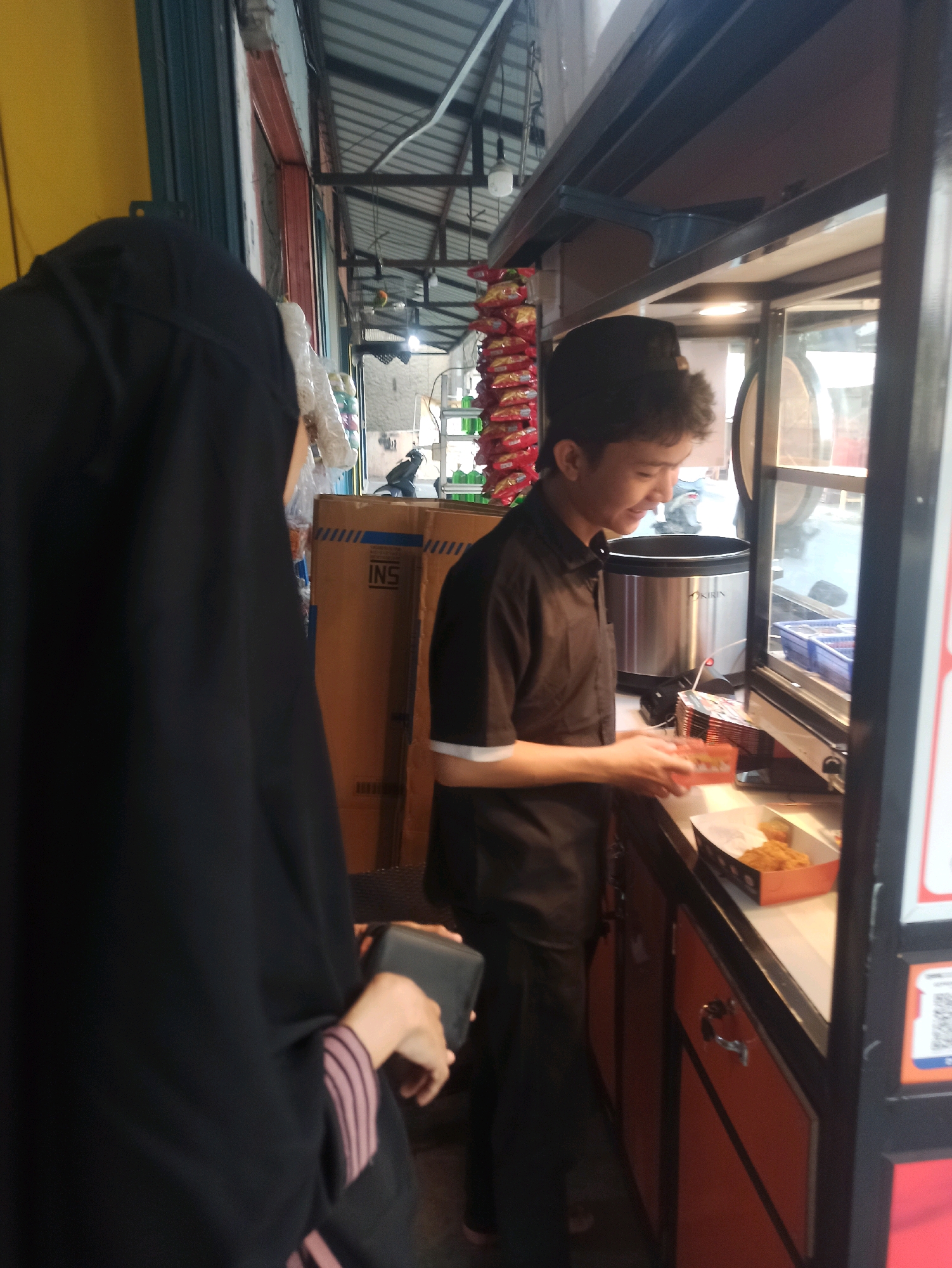 A young male food vendor, appearing to be of Southeast Asian descent and wearing a black collared shirt, smiles as he places a takeaway box of fried chicken onto the counter of his food stall. He is positioned behind the counter, facing the customer. In the foreground, a person wearing a black hijab and a dark, long-sleeved garment with striped cuffs is partially visible, holding a dark rectangular object. The food stall features a glass display case on the right, revealing various food items. Shelves are visible above the counter, stocked with packages of snacks and a large metal pot. The background shows the exterior of shops with corrugated metal roofs, suggesting an outdoor market or street-side setting in Telagamurni, Indonesia. The scene is lit by overhead lights and natural daylight, creating a moderately bright environment.