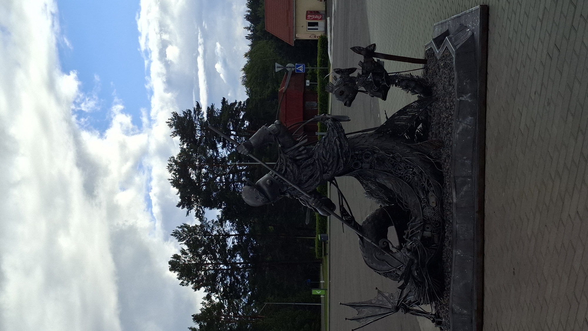 This is a full-shot, eye-level, outdoor photograph of a metal sculpture depicting a medieval warrior in armor, engaged in combat with a dragon. The sculpture is dark gray, intricately detailed, and appears to be made of welded metal pieces. The warrior is shown wielding a spear, thrusting it towards the dragon, which is coiled around the warrior's lower body. The dragon's head is visible near the front of the sculpture, with sharp teeth and scales. Two smaller, fish-like creatures are also part of the sculpture, positioned to the right of the main figures.
The sculpture is situated on a raised platform filled with small, dark stones. The platform itself is placed on a paved area with brick-like patterns. In the background, there are tall, dark green pine trees, suggesting a wooded area. Several buildings are visible behind the trees, including a red-roofed structure and a yellow building with a Coca-Cola sign. Traffic signs, including a blue pedestrian crossing sign, are also present. The sky above is partly cloudy with patches of blue. The scene appears to be a public space, possibly a park or rest stop, located within or near the Асіпавіцкі сельскі Савет in Belarus. There are no people visible in the photograph.