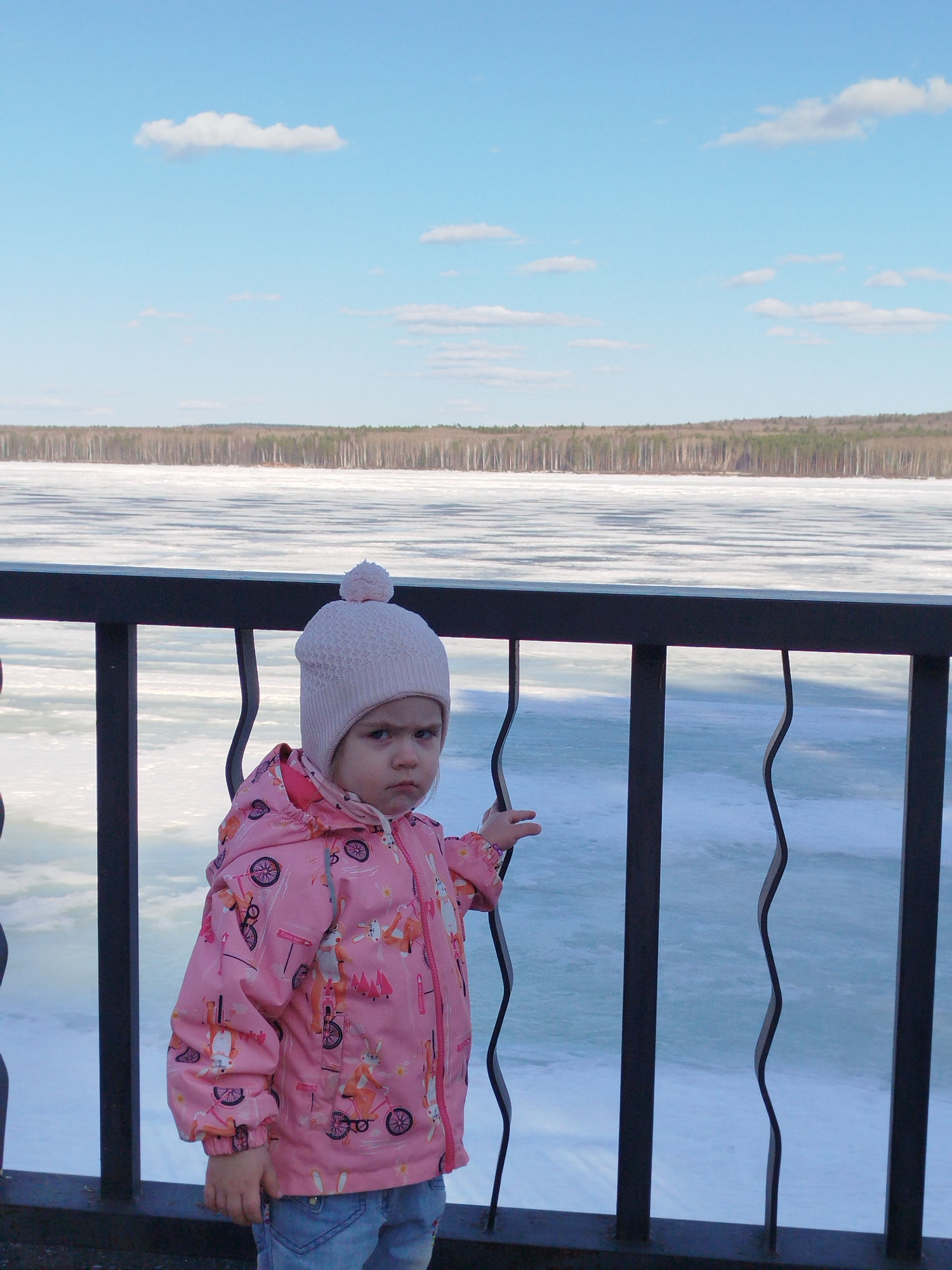 A young child stands by a black metal railing overlooking a frozen lake. The child wears a pink patterned jacket, blue jeans, and a light pink knit hat with a pom-pom. The child appears to be frowning. In the background is a frozen lake and a line of trees. The sky is blue with scattered clouds. Based on the location in the city of Chaikovsky, Russia, this scene depicts the railing overlooking the frozen Kama River.
