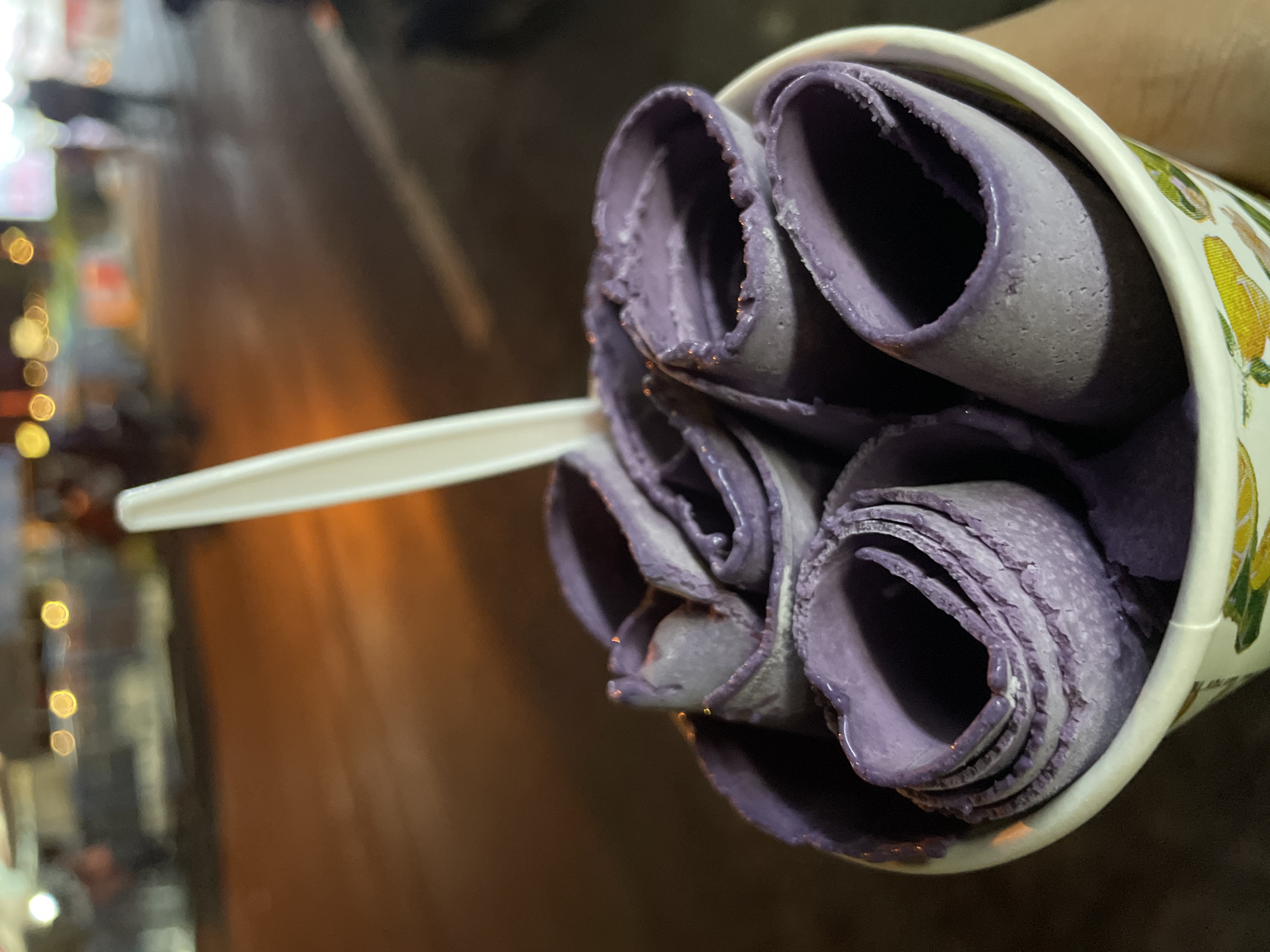 A close-up shot shows a white paper cup filled with rolled ice cream, which is purple in color. A white plastic spoon is inserted into the ice cream. The ice cream is presented in thin, rolled layers, with some liquid appearing to have melted and pooled at the bottom of the cup. The cup has a decorative pattern with images of corn on the cob. The background is blurred, suggesting an outdoor setting at night. Visible elements in the background include streetlights with a warm, orange glow, and what appear to be lights from buildings or vehicles, indicating an urban environment. The overall impression is of a casual street food experience. The location context places this scene in Davao City, Philippines, suggesting a warm climate, and the nighttime setting with artificial lighting is typical of a city environment after dark. No visible text is present on the ice cream or the spoon.