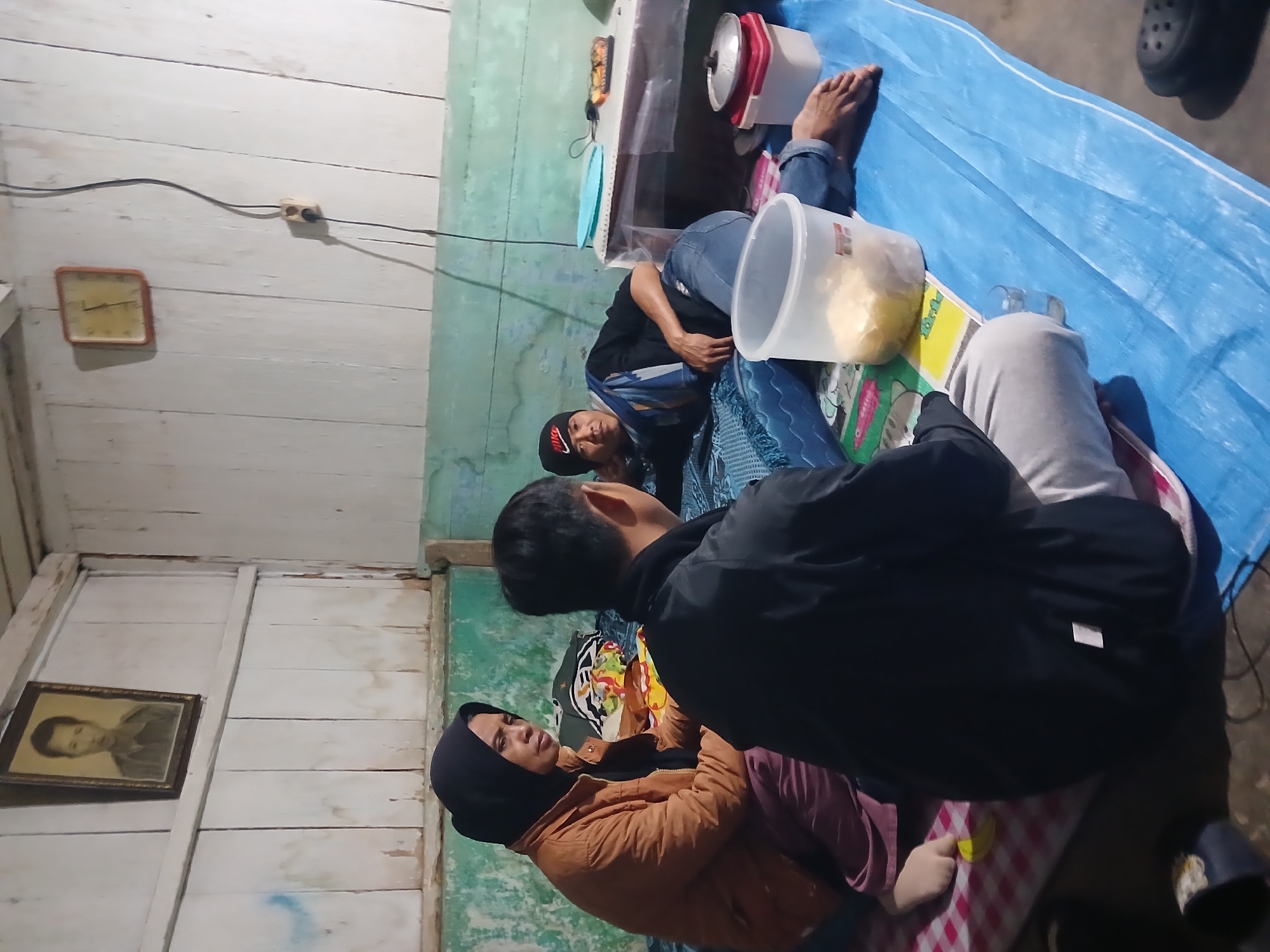 In Candiroto, Indonesia, three individuals are observed in a dimly lit room with wooden walls and ceiling. A framed portrait of a man hangs on the upper left wall, and a square clock is mounted on the right wall. The room is furnished with bedding laid out on the floor, covered by a blue tarp. Two individuals are seated on the bedding, facing away from the camera, while a third individual is reclined on the bedding, looking towards the other two. A transparent plastic container filled with food is positioned in the foreground. Also visible are a metal pot with a lid, a clear plastic cup, and various other objects scattered on the floor. The scene suggests a casual indoor gathering or resting space.