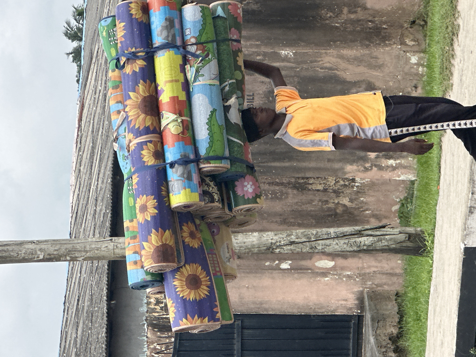 In Badagry, Nigeria, a young man walks carrying a large stack of rolled-up mats on his head. The mats are colorful and patterned, with some featuring sunflowers and others depicting cartoon animals and letters. He is wearing an orange and grey polo shirt and black athletic pants with white stripes. The background shows a rustic, aged building with pinkish-tan walls, a corrugated metal roof, and a wooden utility pole. Lush green grass grows along the side of a concrete pathway. The sky is overcast with a hazy, cloudy appearance.