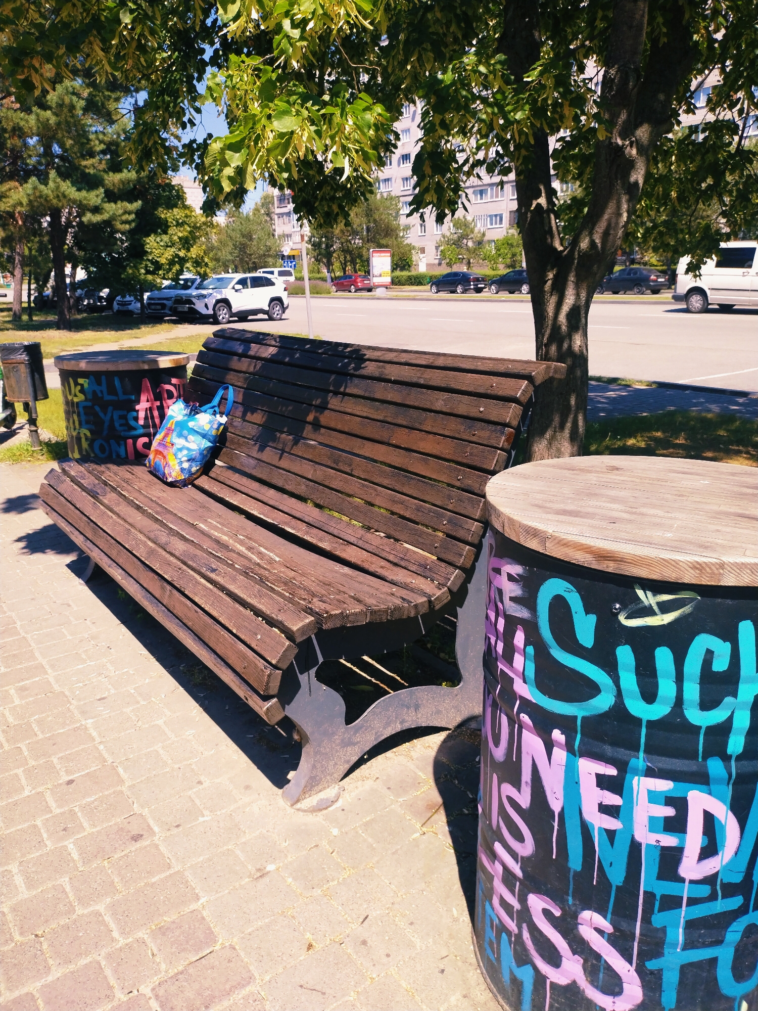 An outdoor scene in Horishni Plavni, Ukraine, features a wooden park bench situated on a paved walkway. Adjacent to the bench are two cylindrical trash receptacles. The left receptacle is a dark metal bin, partially visible. The right receptacle is a black barrel adorned with colorful graffiti. The bench has a blue tote bag with an Eiffel Tower print placed on it. Behind the bench, a tree with lush green foliage casts shadows onto the walkway. In the background, a multi-story apartment building and a street with passing cars are visible. The sky is clear and blue, indicating a sunny day.