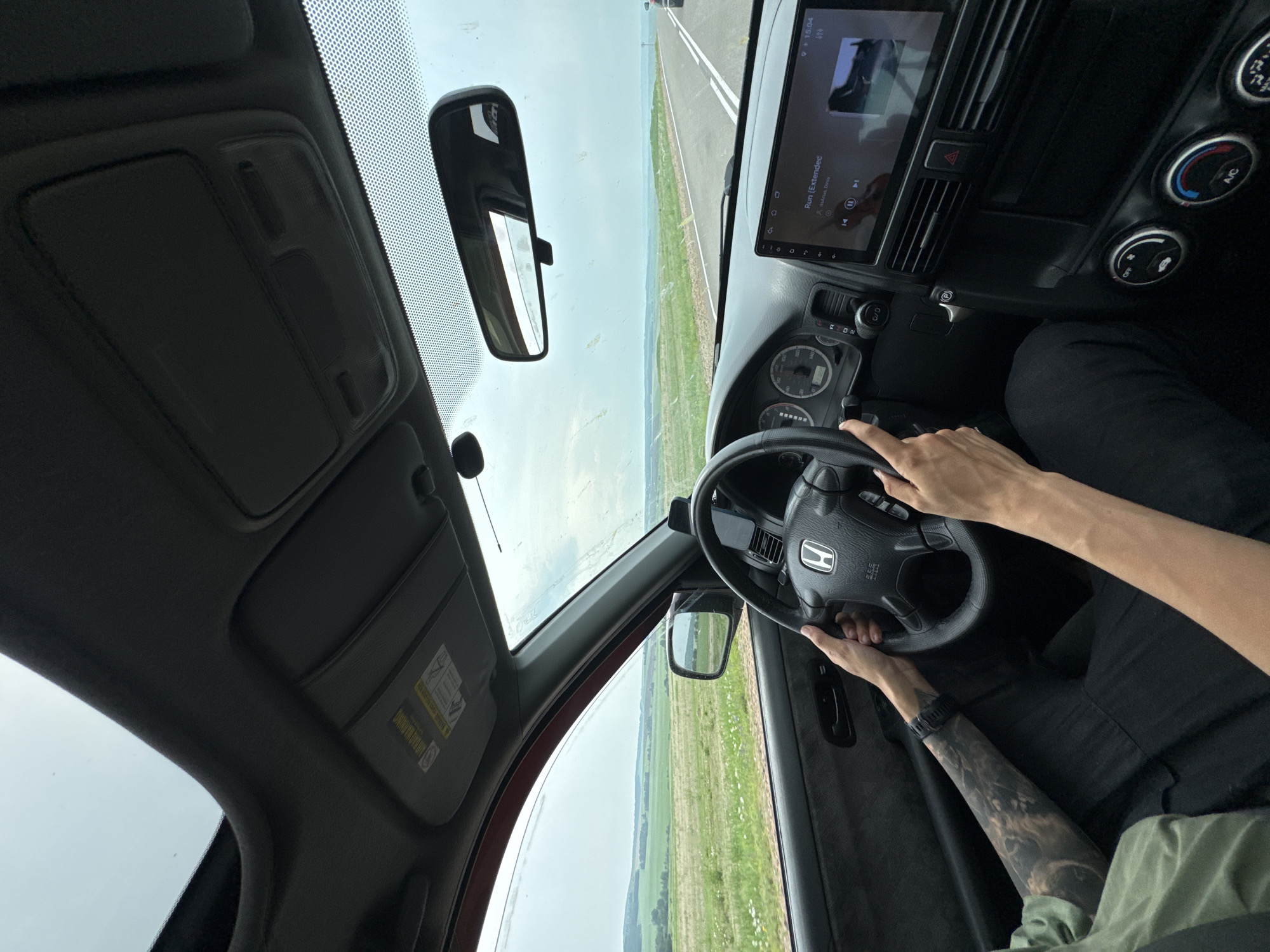 An interior, first-person perspective shot shows a person driving a car on a rural highway. The driver's hands are on the steering wheel, and their left arm, adorned with a tattoo, is visible. The car's dashboard is visible, including the instrument cluster and a touchscreen infotainment system displaying album art and song title. Outside the car, a vast, green landscape stretches to the horizon under an overcast sky. The road ahead appears empty, with white lines marking the lanes. The location is identified as Berezovyi, Russia.