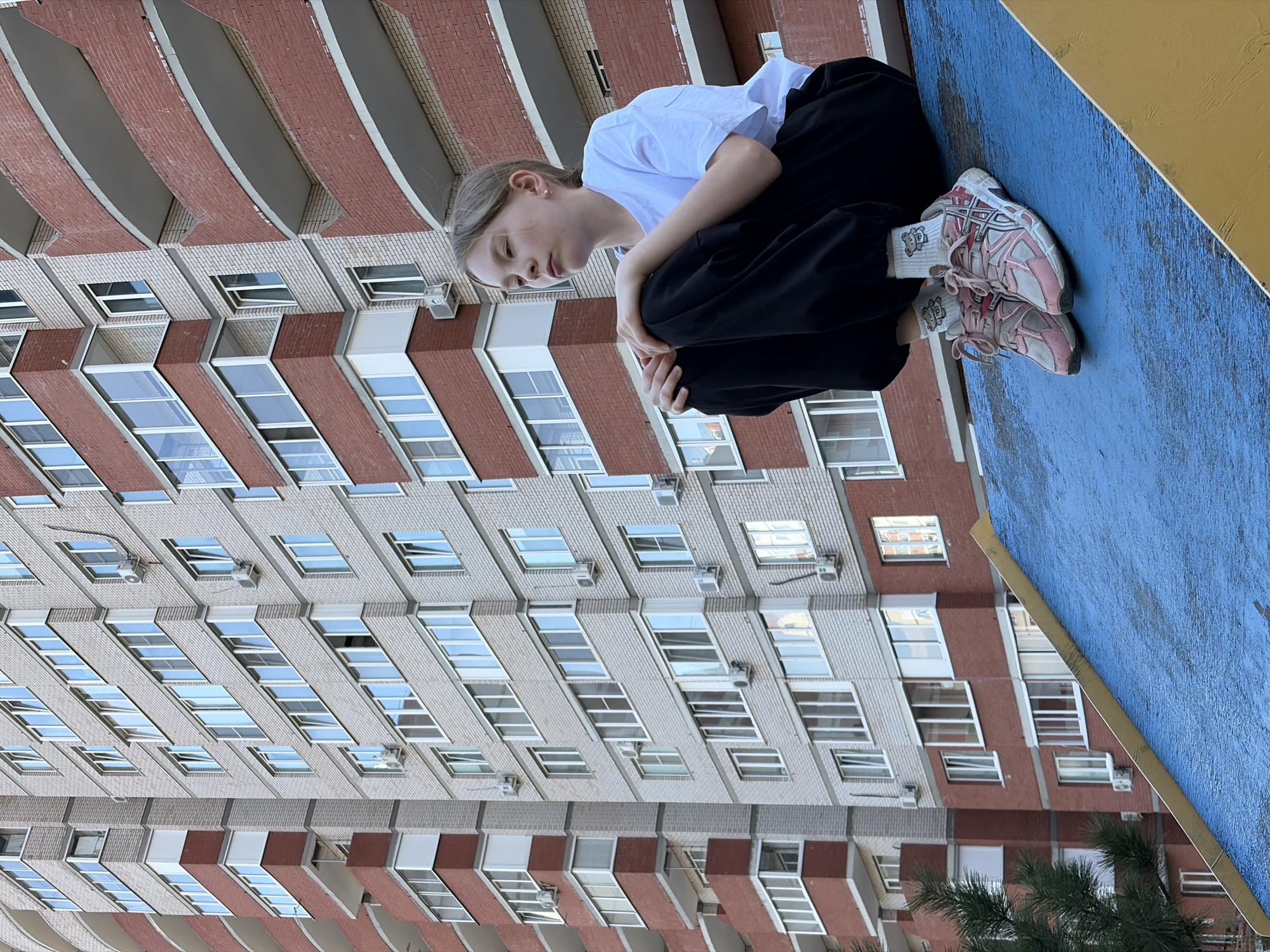 A young girl wearing a white t-shirt and black sweatpants sits with her knees pulled to her chest on a blue and yellow platform. She wears pink and white sneakers and white socks with a pattern. Behind her is a multi-story brick building with many windows, some reflecting the sky. The photo may be taken in Izhevsk, Russia.