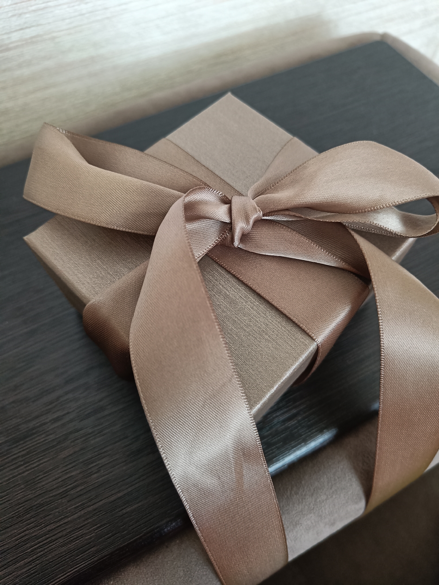 A close-up, angled shot shows a small, square, light brown gift box tied with a matching satin ribbon. The ribbon is knotted in the center of the box, with loops and ends draped over the sides. The box has a subtle textured surface. The background is divided into two parts: the upper left is a light-colored surface with a wood grain pattern, possibly a wall or shelf. The lower right is a dark, textured surface, likely a table or desk. The lighting casts soft shadows, highlighting the sheen of the ribbon and the texture of the box. There are no people visible in the image. The setting does not provide any specific information that would allow for localization to the city of Первомайское, Russia.