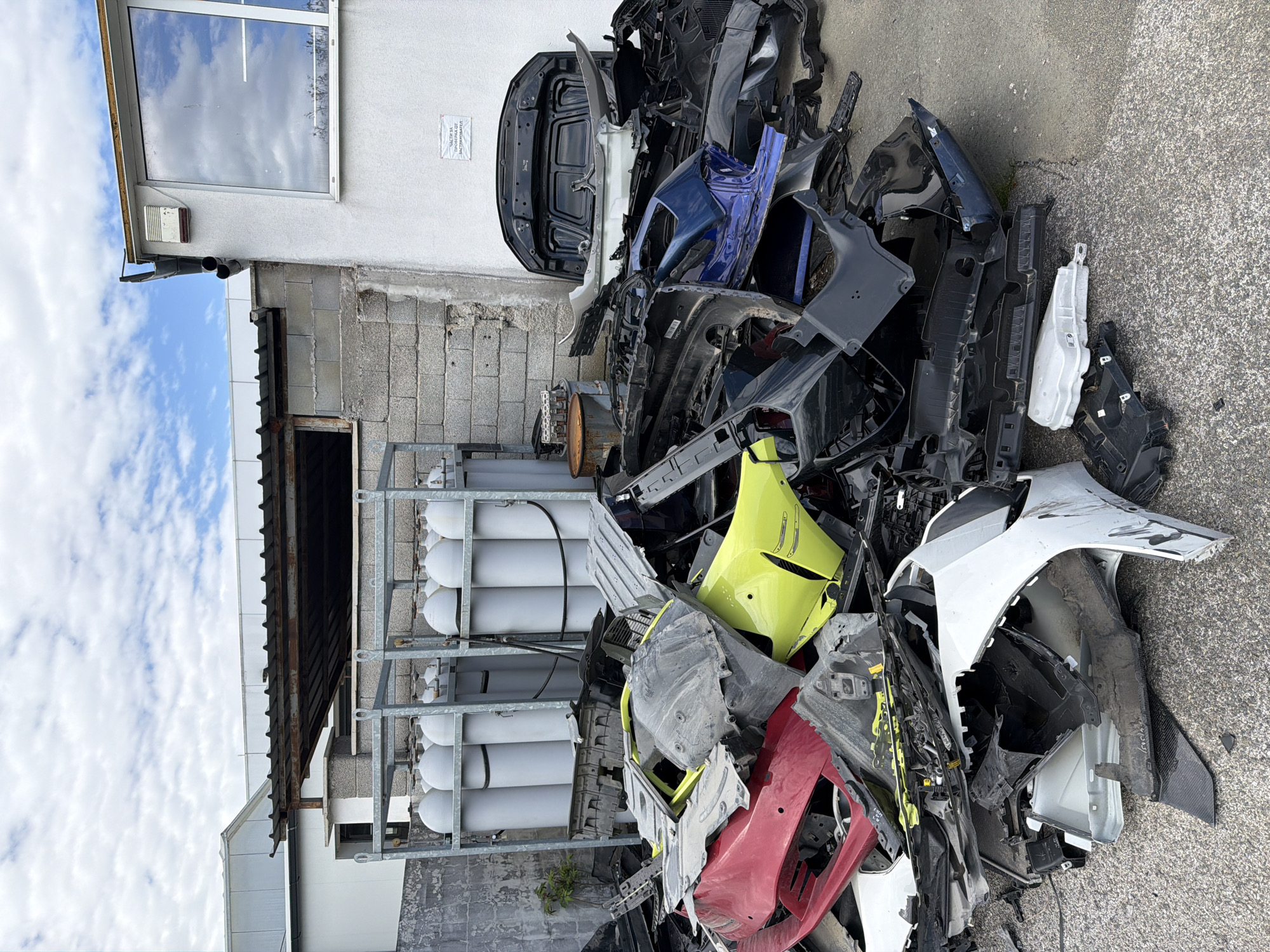 The image shows a pile of discarded car parts next to a building in an urban environment, likely Burgas, Bulgaria. The parts are primarily painted body panels, including bumpers, hoods, and other exterior pieces in various colors such as red, yellow, blue, white, and black. The parts are piled haphazardly on a paved surface, suggesting a scrapyard or auto repair facility. Behind the pile is a building with a window reflecting the sky and clouds. Adjacent to the building is a metal rack holding several white cylindrical tanks. The overall scene conveys a sense of automotive waste and disrepair.