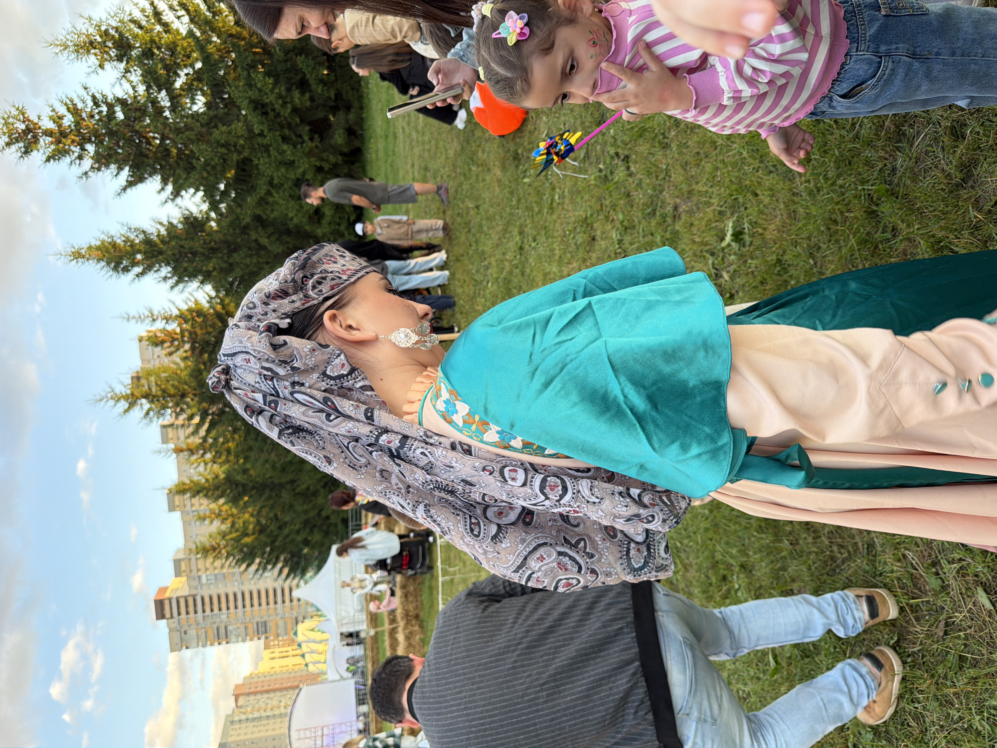 In an outdoor setting with a grassy field, a woman wearing a traditional patterned headscarf and a teal and peach outfit stands on the right side of the frame, facing downwards. She is adorned with large, silver earrings. To her right, a young girl in a pink and white striped shirt and blue jeans looks to her right, holding a colorful pinwheel. In the background, several evergreen trees and apartment buildings are visible under a sky with scattered clouds. Other individuals and tents are present in the middle ground. On the left side of the frame, a person in a dark, striped jacket and light blue jeans is seen from the back, walking away from the camera.