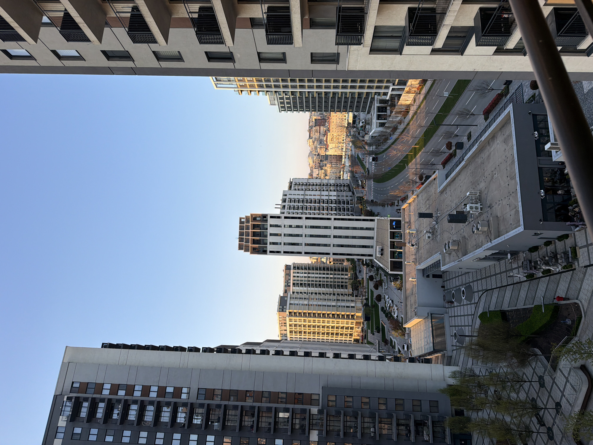 A high-angle daytime shot reveals a cityscape, likely Belgrade, Serbia, judging by the modern architecture style of Belgrade Waterfront. The image is framed by tall apartment buildings in the foreground and top. A wide street with light traffic is seen stretching into the distance, flanked by more modern buildings. The overall lighting suggests late afternoon with the sun casting shadows. The image shows an urban environment in Belgrade.