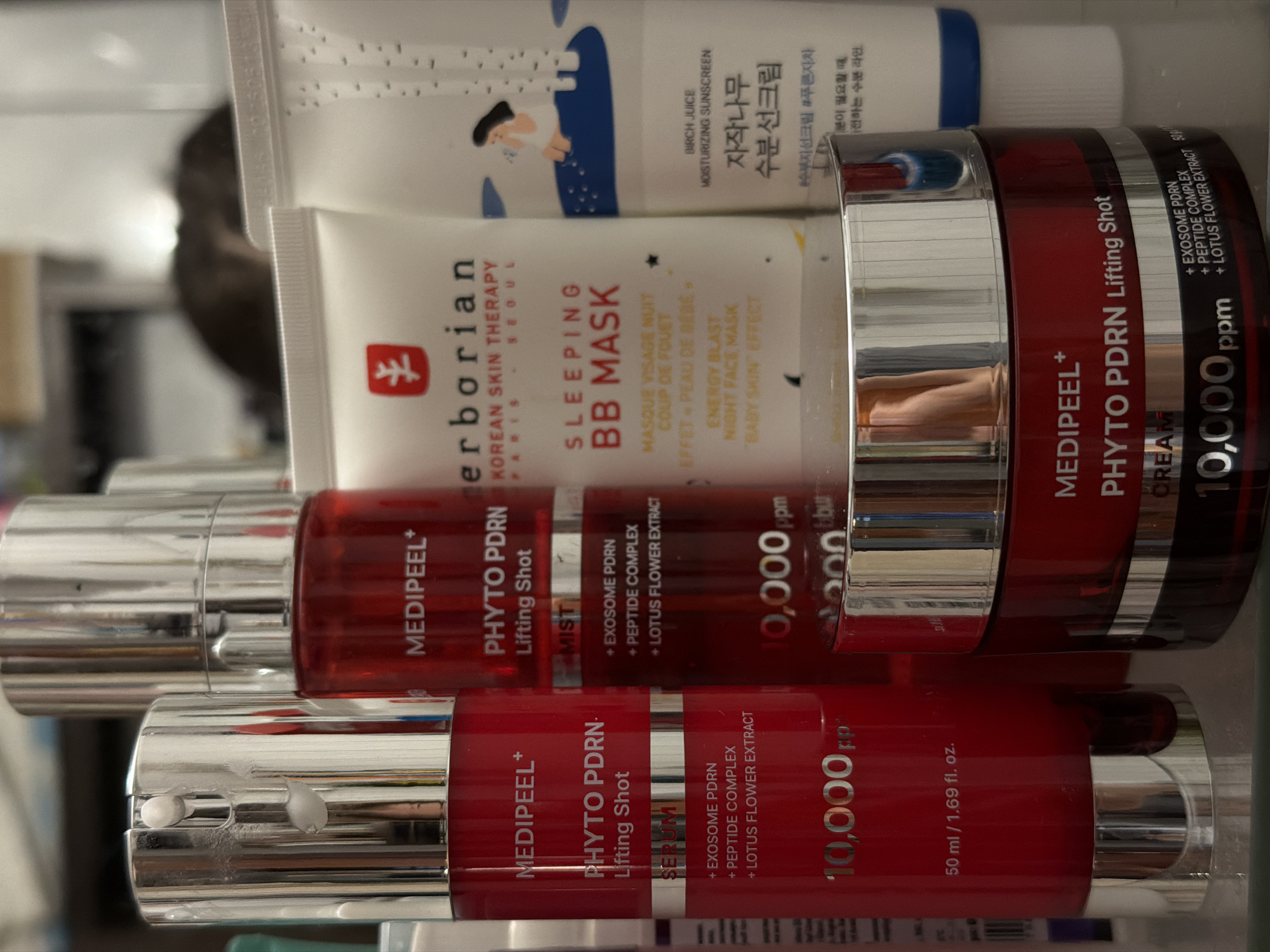 The scene is located in the city of Khimki, Russia. Five cosmetic products are arranged on a reflective surface, likely glass or polished material, reflecting the lower portions of the containers.
In the left foreground, a red cylindrical pump bottle with a silver dispenser top is visible. It is labeled "MEDIPEEL+ PHYTO PDRN Lifting Shot SERUM" and specifies "10,000 ppm" with active ingredients including "+ EXOSOME PDRN", "+ PEPTIDE COMPLEX", and "+ LOTUS FLOWER EXTRACT". The volume is indicated as "50 ml / 1.69 fl. oz.". White product residue is present on the silver pump mechanism.
Directly behind this serum bottle, a similar red cylindrical bottle with a silver spray top is positioned. This product is labeled "MEDIPEEL+ PHYTO PDRN Lifting Shot MIST" and lists the same active ingredients and "10,000 ppm" concentration.
To the right of the central Medipeel products, in the foreground, a red jar with a wide silver lid is displayed. It is labeled "MEDIPEEL+ PHYTO PDRN Lifting Shot CREAM" and also indicates "10,000 ppm" of "+ EXOSOME PDRN", "+ PEPTIDE COMPLEX", and "+ LOTUS FLOWER EXTRACT".
Behind the Medipeel cream jar and slightly to the right, a white squeeze tube is visible. It features a red, stylized tree logo and the brand name "erborian", with text "KOREAN SKIN THERAPY PARIS SEOUL". The product is identified as "SLEEPING BB MASK", with additional descriptions such as "MASQUE VISAGE NUIT", "COUP DE FOUET", "EFFET "PEAU DE BEBE"", "ENERGY BLAST", "NIGHT FACE MASK", and ""BABY SKIN" EFFECT".
To the far right of the image, partially obscured, is another white squeeze tube. This tube features a blue illustration depicting a figure beside birch trees and is labeled "BIRCH JUICE MOISTURIZING SUNSCREEN", with Korean text "자작나무 수분선크림".
The background is significantly out of focus, revealing a blurred, dark, amorphous shape consistent with a resting animal, possibly a cat or dog, behind the products. The overall setting is an indoor environment with ambient lighting.