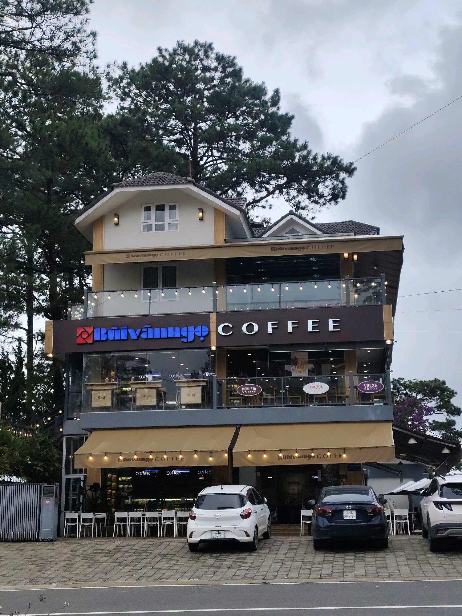 The image captures a three-story building housing a coffee shop in Da Lat, Vietnam. The building features a modern design with glass facades and balconies decorated with string lights. The ground floor appears to be the main coffee shop area, with tables and chairs visible through the windows. A prominent sign displays "Bùi Văn Ngô COFFEE" in large letters. The upper floors also have windows and balconies, with awnings extending over the lower levels.
Outside the coffee shop, several cars are parked on a paved area in front of the building. Two white cars and a dark blue sedan are visible. The sedan has a Vietnamese license plate. White chairs are neatly arranged outside the entrance to the coffee shop, suggesting an outdoor seating area. Tall pine trees surround the building, characteristic of Da Lat's landscape. The sky is overcast, with a few wispy clouds. The overall scene depicts a commercial establishment in a natural, hilly environment.