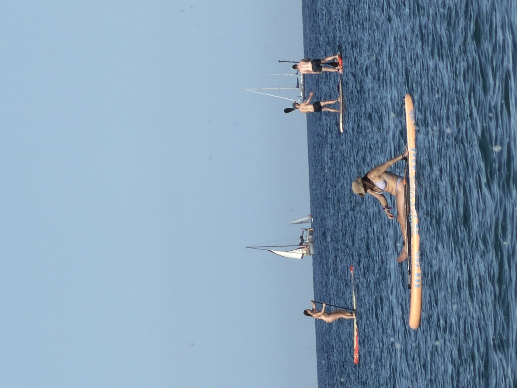 The image shows a group of people paddleboarding on a body of water with sailboats in the background. In the foreground, a woman in a bikini and a hat is sitting on an orange paddleboard. To her right, a man in black shorts is standing on a paddleboard, facing away from the camera. To the right of him, another man in black shorts is standing on a paddleboard, also facing away from the camera. In the background, a woman in a bikini is standing on a paddleboard and holding a paddle. Several sailboats are visible on the water. The sky is clear and blue. The scene appears to be taking place in a coastal area, possibly near a city given the sailboats and the overall recreational activity. Without specific landmarks, it is impossible to definitively place this scene within Barcelona, Spain.