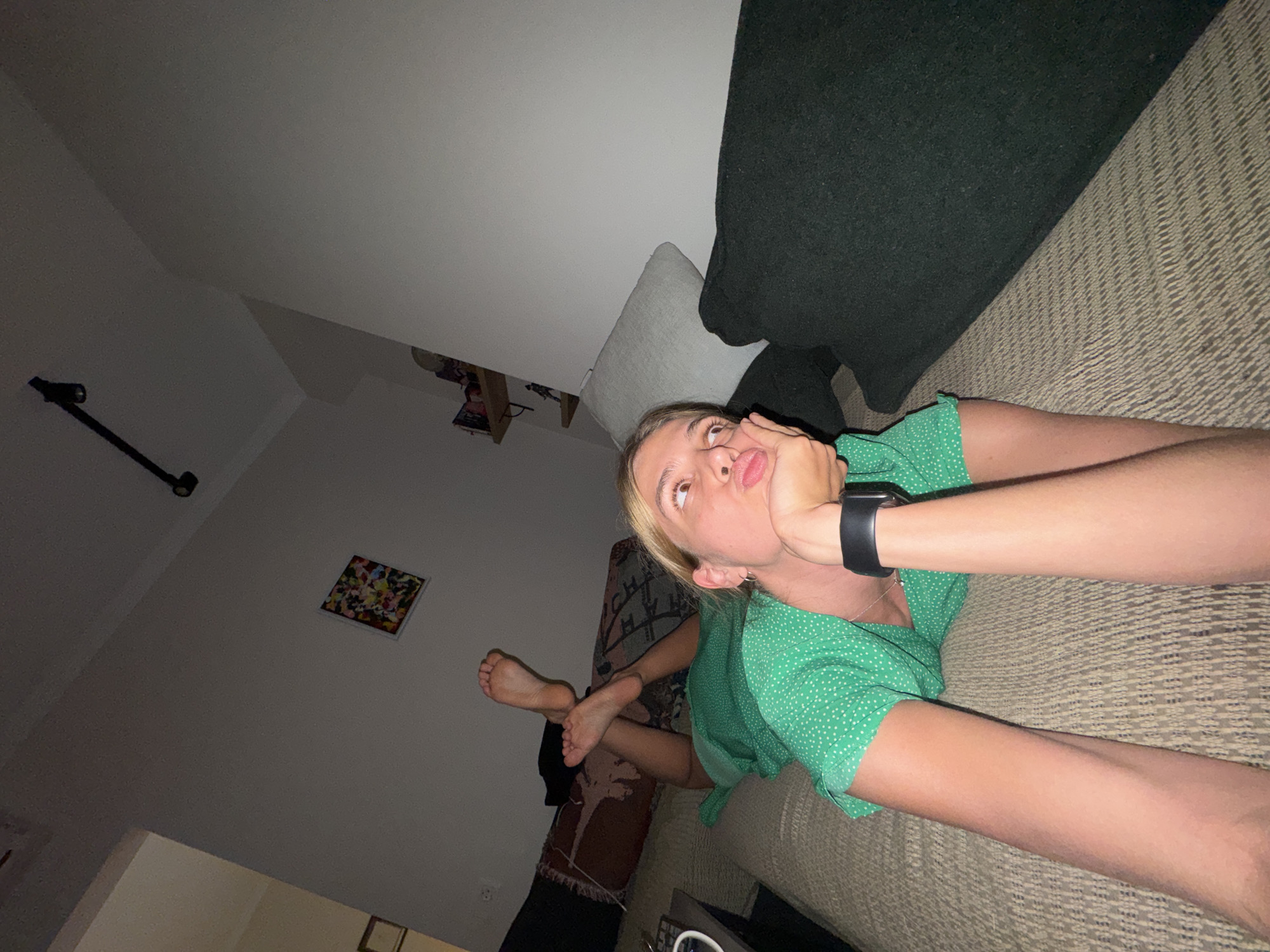 A low-angle, indoor photograph captures a young woman lying in a relaxed pose on a textured, light-brown upholstered surface, likely a couch. She is wearing a green dress with white polka dots and a dark smartwatch on her left wrist. Her blonde hair is pulled back, and she looks upwards with a thoughtful or playful expression, her mouth slightly pursed and her right hand resting near her chin. Her bare feet are visible in the mid-left, crossed in a relaxed manner.
Behind the woman, a dark green cushion and a lighter-colored cushion are visible on the upholstered surface. The background consists of a light-colored wall with an angled ceiling, suggesting a unique architectural space, possibly an attic or a room with sloped ceilings. A dark, stick-like fixture, potentially a light or camera mount, is attached to the wall in the upper left. Further along the wall, a small framed picture with colorful, abstract elements is visible.
The lighting in the image is dim, indicating an indoor setting, likely evening or night. The overall atmosphere conveys relaxation and informality. No text is visible in the image.