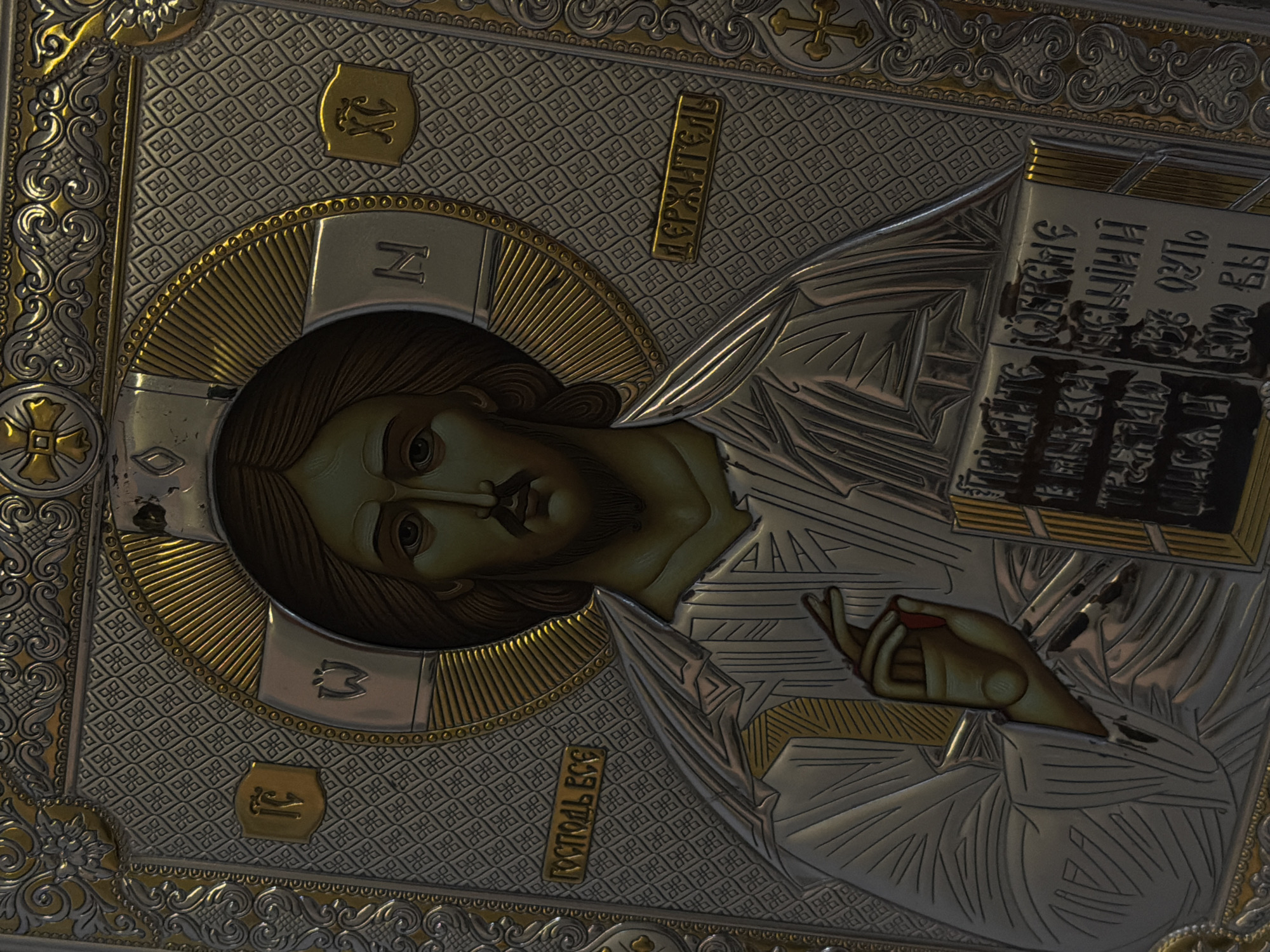 This image captures a close-up, vertically oriented view of an ornate Orthodox icon of Jesus Christ, known as the Pantocrator. The icon features a metallic riza (oklad) made of silver and gold-toned metals, covering a painted image of Christ's face and hands.
The central figure is Jesus Christ, depicted with a serene yet somewhat somber expression, dark hair, beard, and mustache. His face is painted in a traditional Byzantine style, with large, observant eyes. A golden, rayed halo encircles his head, containing the Greek letters "Ο Ω Ν" (Omicron, Omega, Nu), signifying "The Being One" or "He Who Is."
Christ's right hand is raised in a traditional gesture of blessing, with two fingers extended and three joined, symbolizing the Trinity and His two natures (divine and human). His left hand holds an open Gospel book, detailed with metallic covers and raised scriptural text. The book displays various lines of Church Slavonic script, including legible words such as "ІИСУСЪ ХРИСТОСЪ" (Jesus Christ) and "ЕСТЬ" (is), suggesting a passage from the Gospels.
The metallic riza is highly detailed with intricate relief patterns. The background features a textured pattern of repeating squares, interspersed with larger floral and scrollwork designs, all in silver and gold tones. Small gold crosses are visible in the upper corners of the icon's frame.
Several vertical text plaques are incorporated into the riza:
- To the viewer's left of Christ's head, the Cyrillic word "ДЕРЖИТЕЛЬ" (Derzhitiel), meaning "Sustainer" or "Pantocrator."
- To the viewer's right of Christ's body, the text "ИСТОЧНИКЪ ВСЕ" (Istochnik Vse), meaning "Source of All."
- Near the halo, the abbreviated Greek letters "IC" and "XC" are visible, standing for "Іисусъ Христосъ" (Jesus Christ).
The icon's metallic surface reflects light, creating highlights and shadows that emphasize the three-dimensional quality of the relief work. The image's overall mood is one of spiritual reverence and historical craftsmanship. The location context provided is Sofiivska Borshchahivka, Ukraine.