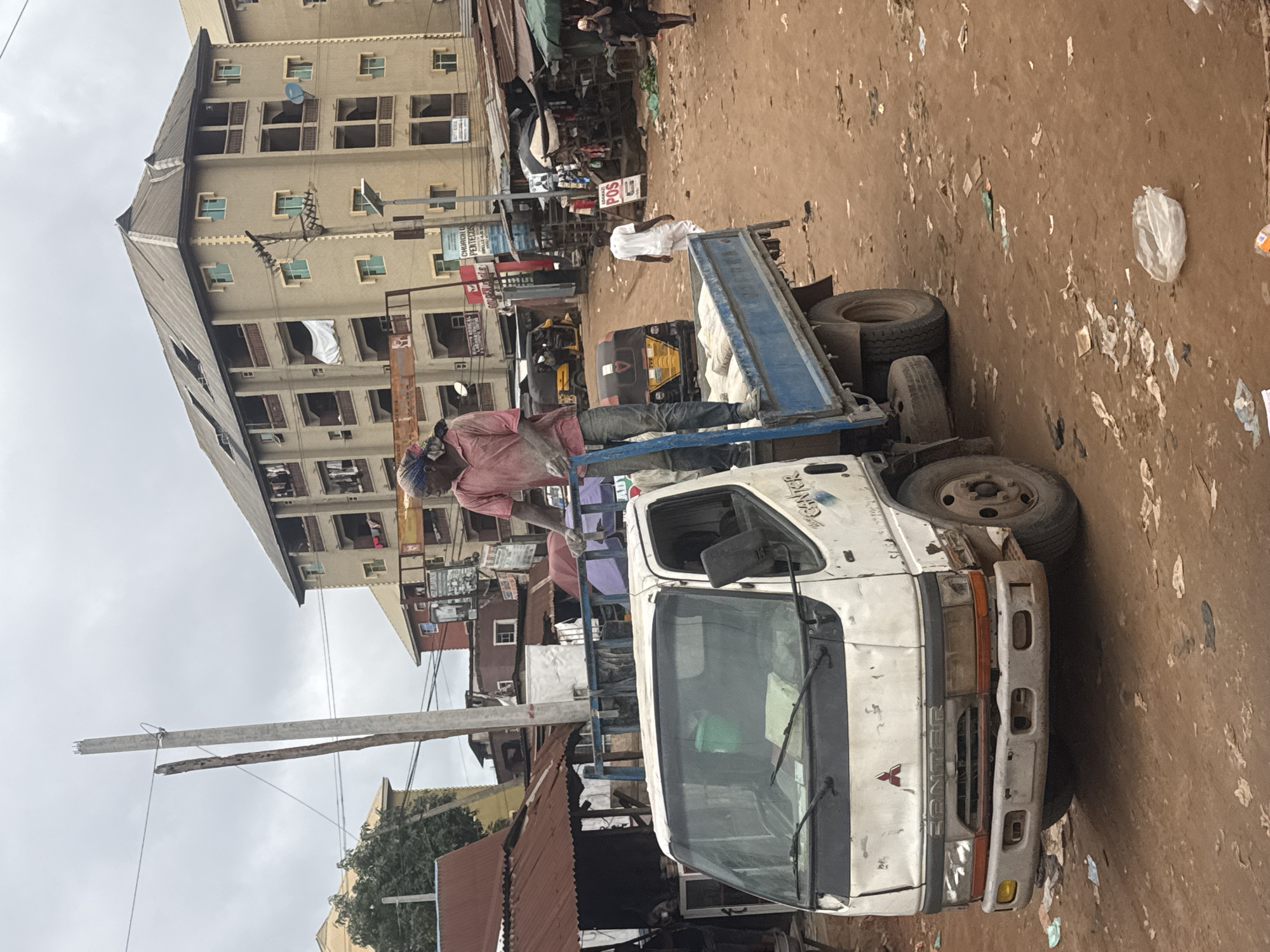 A man wearing a pink shirt and blue jeans stands on the back of a white flatbed truck, holding onto a blue rail. The truck is loaded with bags. The man is dusty, and his face is partially obscured by a headscarf. The truck is parked on a dirt road in what appears to be a busy market area in Nkpor, Nigeria. In the background, there are multi-story buildings, smaller market stalls with corrugated metal roofs, and other vehicles, including a yellow auto-rickshaw. The ground is covered in dirt and scattered debris, suggesting an unpaved and possibly unkempt environment. The sky is overcast.