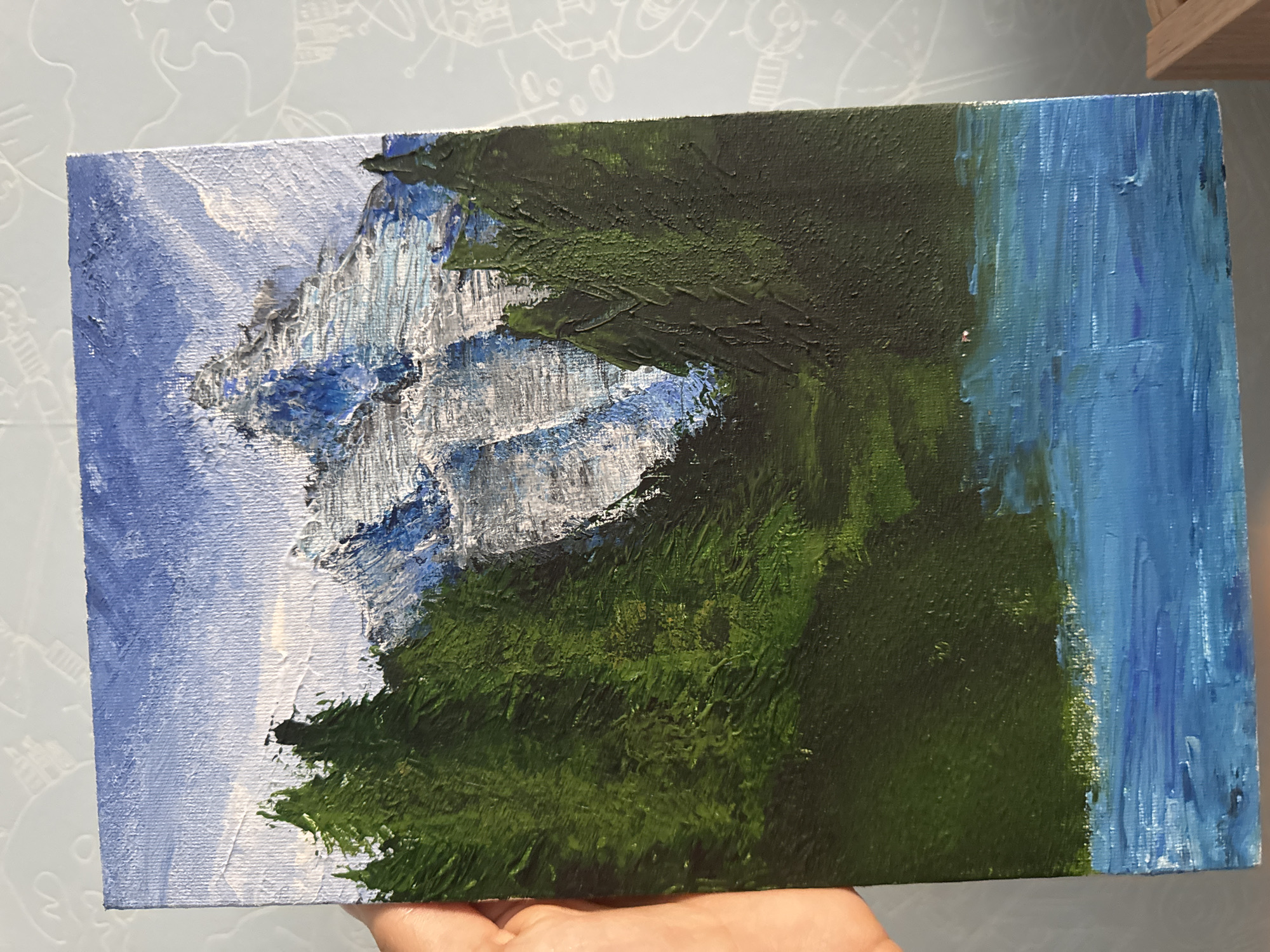 A vertical acrylic painting on canvas depicts a mountain landscape. In the foreground, a body of water with visible brushstrokes in shades of blue and white reflects the sky. Behind the water, a dense forest of dark green trees occupies the middle ground. Rising above the trees is a snow-capped mountain, rendered in strokes of white, blue, and gray, with sharp, rugged peaks. The sky above is a bright blue with some wispy white clouds. The painting is held by a hand on the left side, partially obscuring the canvas. The background of the photograph is a wall with a faint patterned texture.
