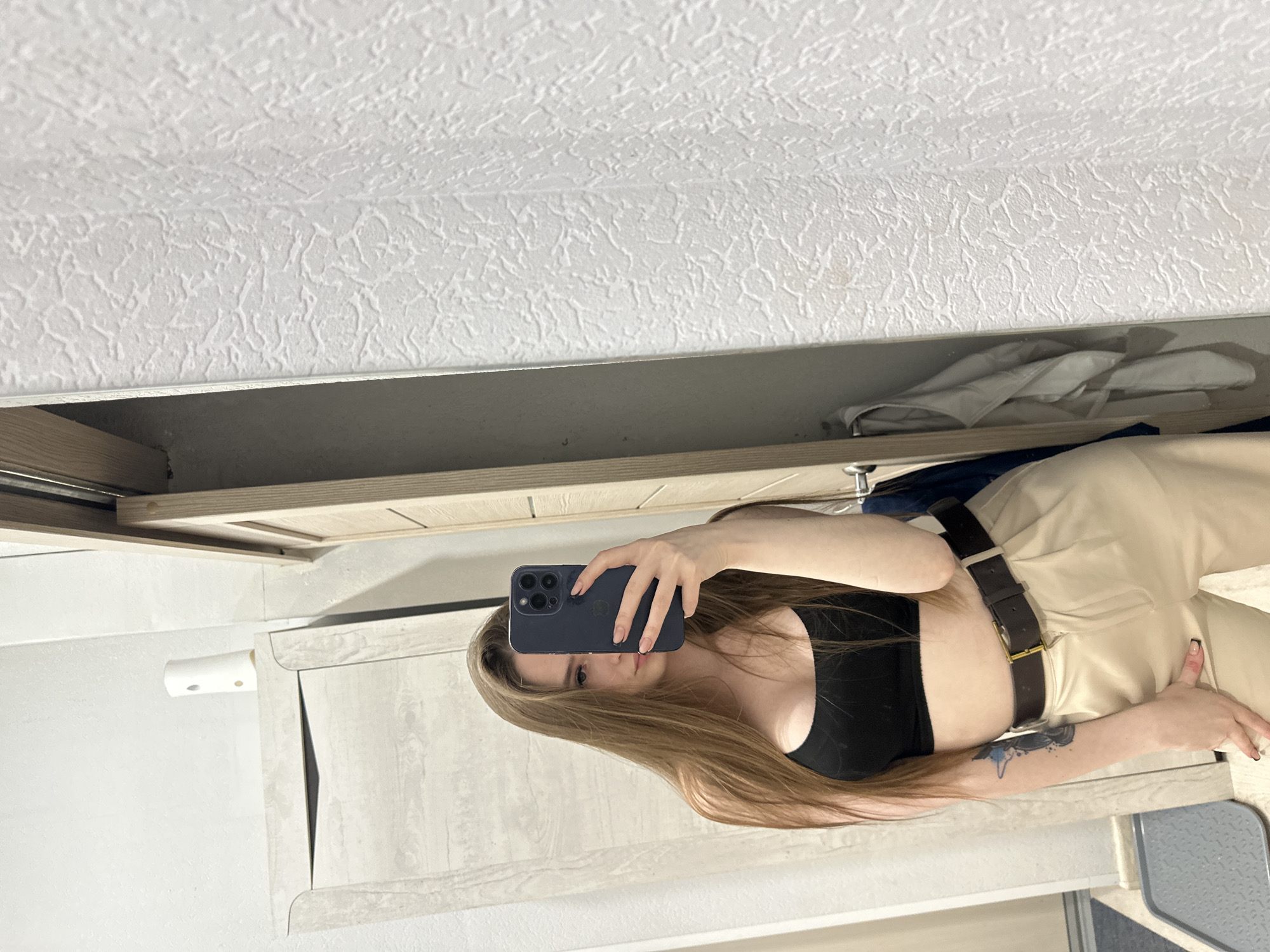 A woman takes a selfie in front of a mirror indoors. She wears a black top and beige pants with a brown belt. Her long, light brown hair is down. The background includes a white textured ceiling and light-colored cabinets. Unknown, Unknown is not identifiable in the image.