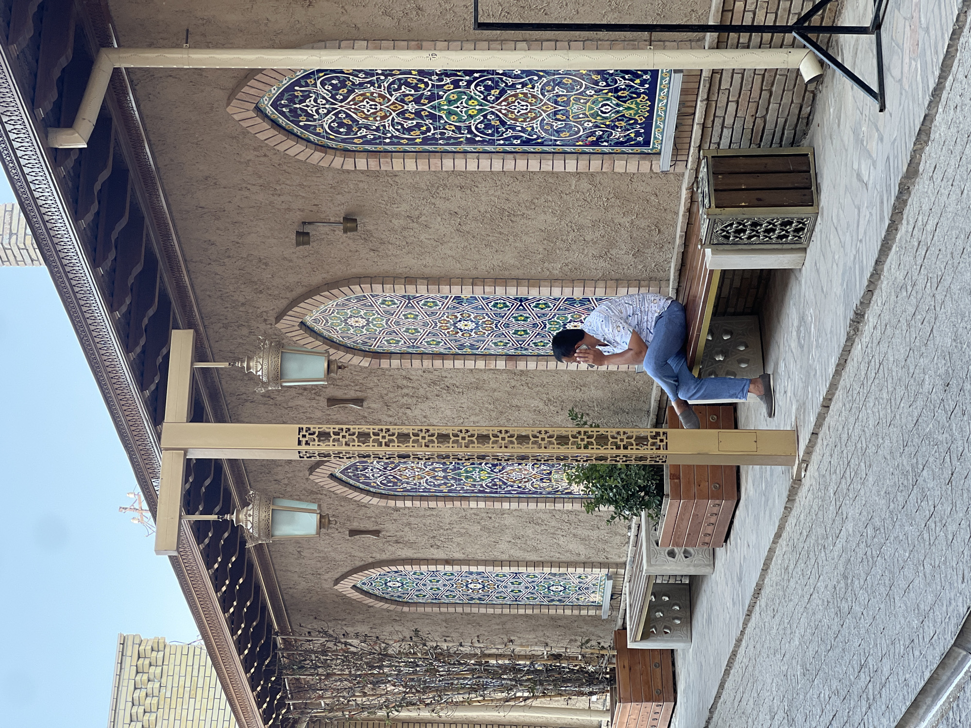 A man is sitting on a bench in front of a building with ornate tilework. He is wearing a light-colored short-sleeved shirt and blue jeans, and he is holding his hand to his face. The building has a textured brown facade with arched windows filled with intricate blue, green, and white tile mosaics. Lamps hang from the building's facade. There are planters with greenery and dry branches, and other benches along the wall. The ground is paved with cobblestones. The scene appears to be in a public outdoor space, possibly a courtyard or a street in Tashkent, Uzbekistan, given the architectural style and the presence of similar decorative elements in the city.