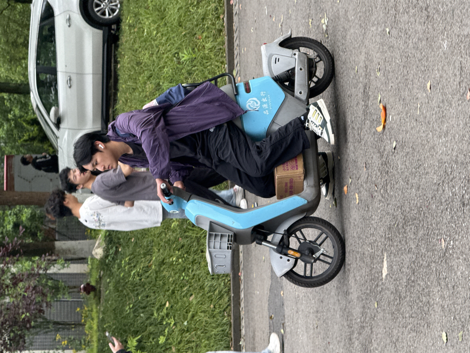 An image captures a street scene, likely in Jiangjin, China, during daylight hours. The central focus is a young man with dark hair, wearing a dark purple jacket over a dark top, dark trousers, and white sneakers with green stripes. He has white earbuds in his ears and is intently looking down and leaning over a light blue and grey electric scooter. A dark backpack rests on the scooter's seat. He appears to be either adjusting something on the scooter or waiting. A brown cardboard box is visible tucked under the scooter's seat/footrest.
The scooter itself is light blue and grey, featuring a small basket-like attachment near the front wheel. On the blue body of the scooter, the Chinese characters "品源出行" (Pǐnyuán Chūxíng, meaning "Pinyuan Travel/Rideshare") and "PINYEAN" in English are visible, indicating it is likely part of a shared mobility service.
In the background, on a grassy verge, two other young men are standing. One wears a grey t-shirt, and the other a white t-shirt, appearing to be conversing or waiting. Further back, a white SUV is parked partially on the grass. The ground is a grey asphalt road, with scattered dry leaves, some appearing orange. The overall lighting suggests an overcast day or perhaps early morning/late afternoon, with no harsh shadows. Green grass and some foliage line the side of the road. A hand holding a phone is partially visible in the lower right corner.