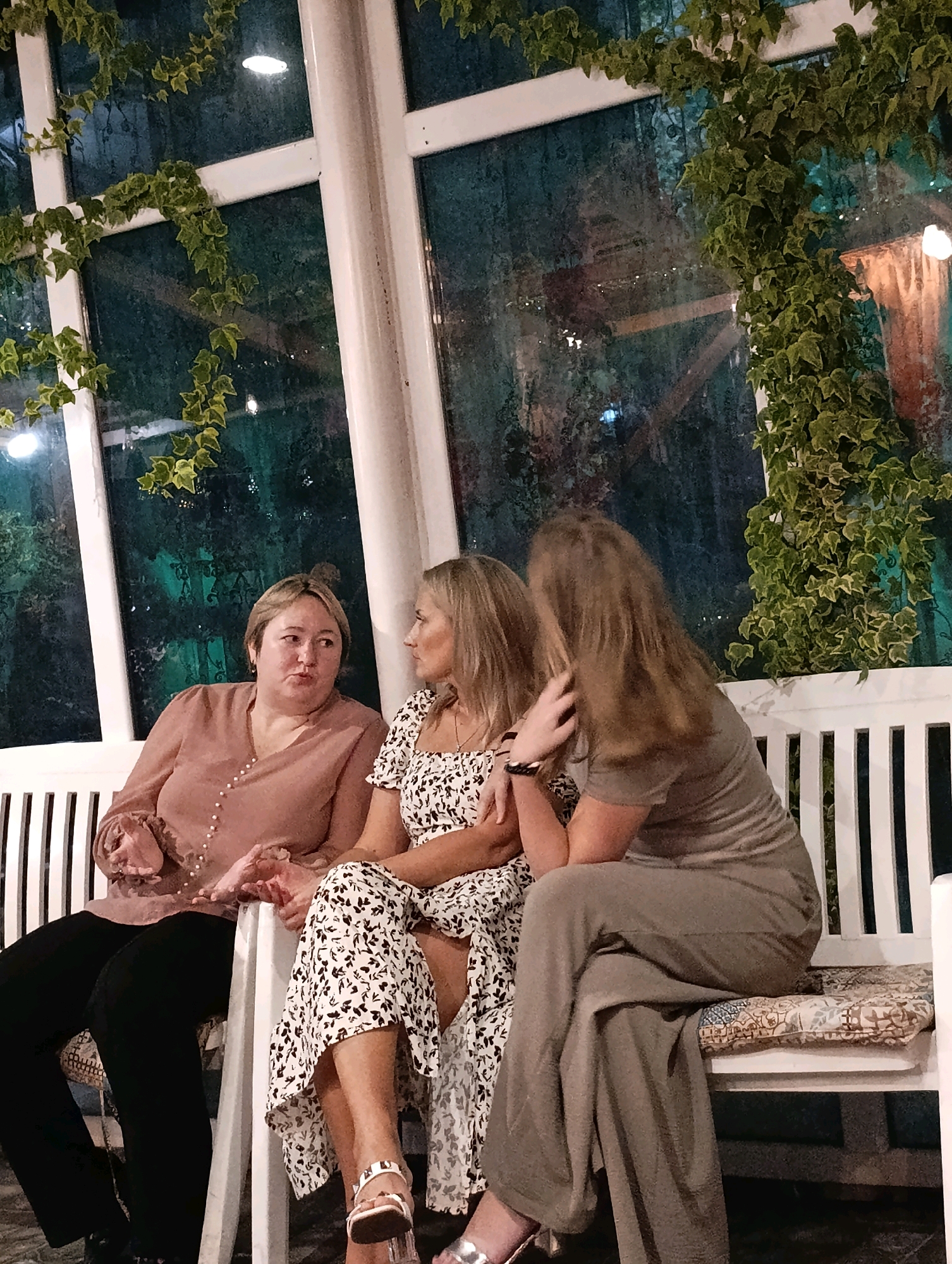 Three women are seated on a white slatted bench, likely outdoors given the greenery draped over a large window or glass partition behind them. The woman on the left, with short blonde hair, wears a muted rose-colored blouse with pearl buttons and black pants. She is looking away from the camera with her hands clasped. The woman in the middle has blonde hair and wears a white dress with a black floral pattern. She is turned towards the woman on the right. The woman on the right, with long brown hair, wears a muted grey outfit and faces the middle woman, her left arm resting on the bench. The environment appears to be a restaurant or cafe with a view of foliage and subtle interior lighting reflected in the glass. The scene is likely within Korzhevo, Moldova.