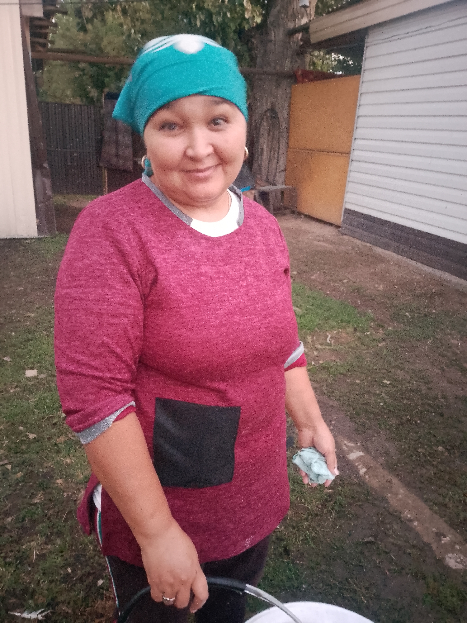 In the village of Keldemurat, Kazakhstan, a woman stands outdoors in what appears to be a yard. She is wearing a turquoise headscarf with a white pattern and a maroon long-sleeved shirt with a white undershirt peeking out. The shirt has a black patch pocket on the front and grey cuffs. She is holding a light blue cloth in her right hand and appears to be holding a white bucket with her left hand, which is mostly out of frame. The ground is covered with grass and some bare, damp soil. In the background, there is a tall tree, a wooden fence, a shed with a white corrugated metal siding, and a large wooden structure. The lighting suggests it is daytime.