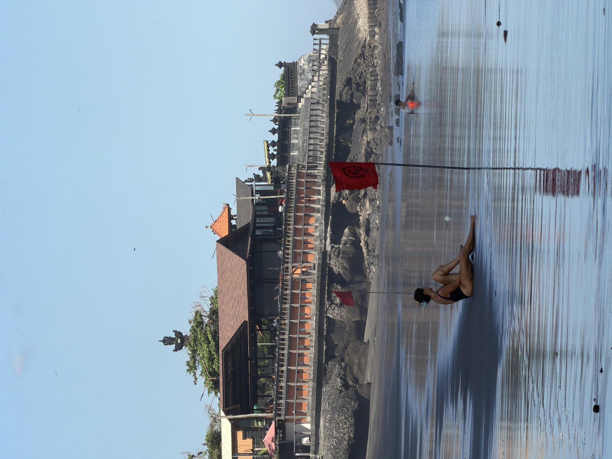 In North Kuta, Indonesia, a woman in a black swimsuit sits on a wet, sandy beach facing left, her body angled away from the camera. Her left leg is bent and positioned in front of her, while her right leg is extended. She is looking towards the water. In the shallow water, another person, wearing an orange garment, is seated. A red flag on a pole stands in the foreground, partially obscuring the seated woman. In the background, a complex of buildings with brown roofs sits on a raised, rocky embankment. A wooden railing runs along the front of the embankment. The sky is clear and blue, with a few distant birds visible. The water reflects the sky and the surrounding structures.