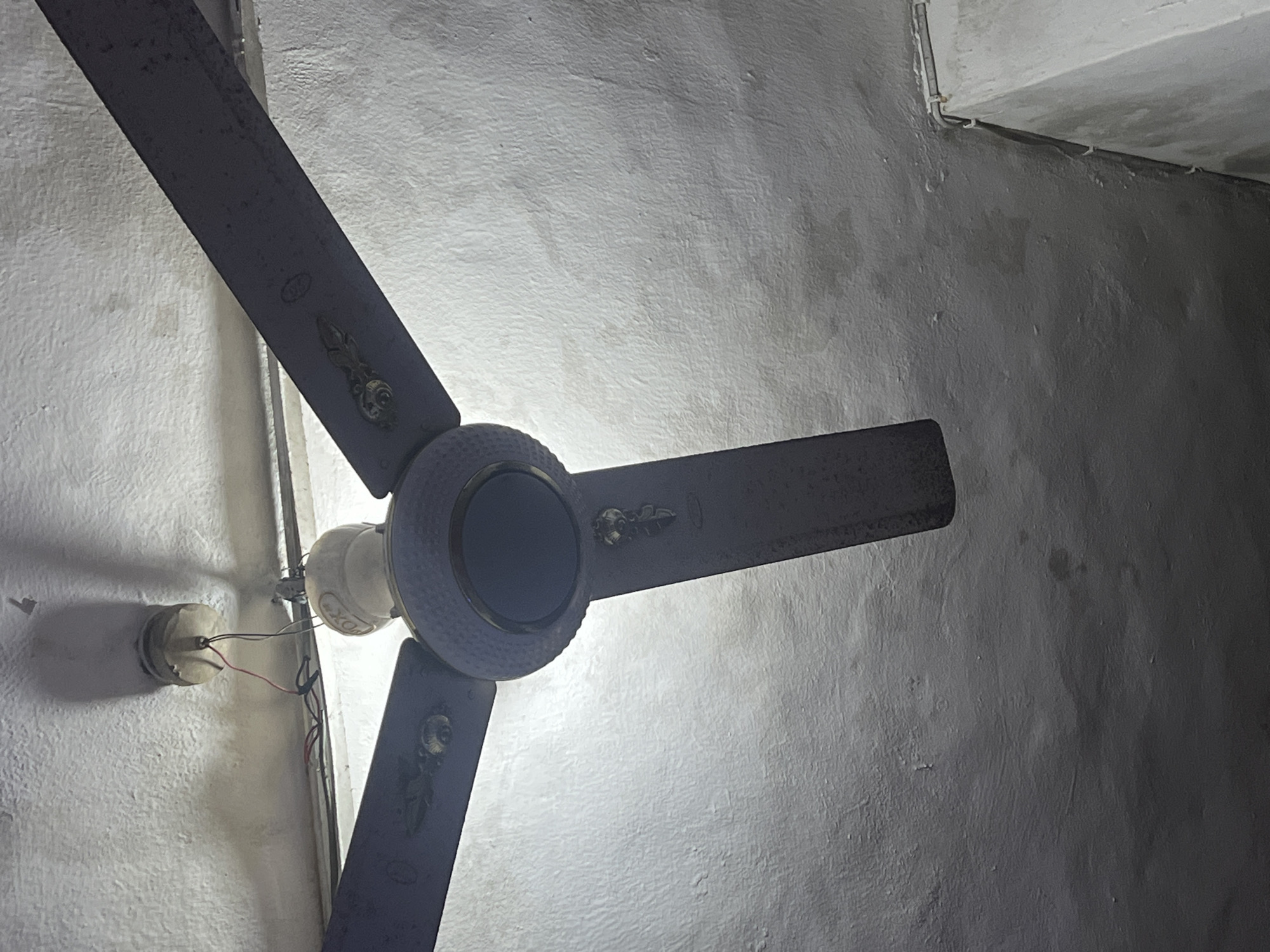 The image shows a ceiling fan with dark-colored blades attached to a white ceiling. The fan's central housing is a mix of white and dark blue, with decorative metallic accents on the blades and housing. Exposed wires are visible connecting to the fan's motor from the ceiling. The ceiling itself is textured and appears to be slightly stained or discolored in places. The lighting in the scene is somewhat dim, casting shadows from the fan onto the ceiling. The angle of the shot is looking upwards at the fan and ceiling. The location is in Ifite-Awka, Nigeria.