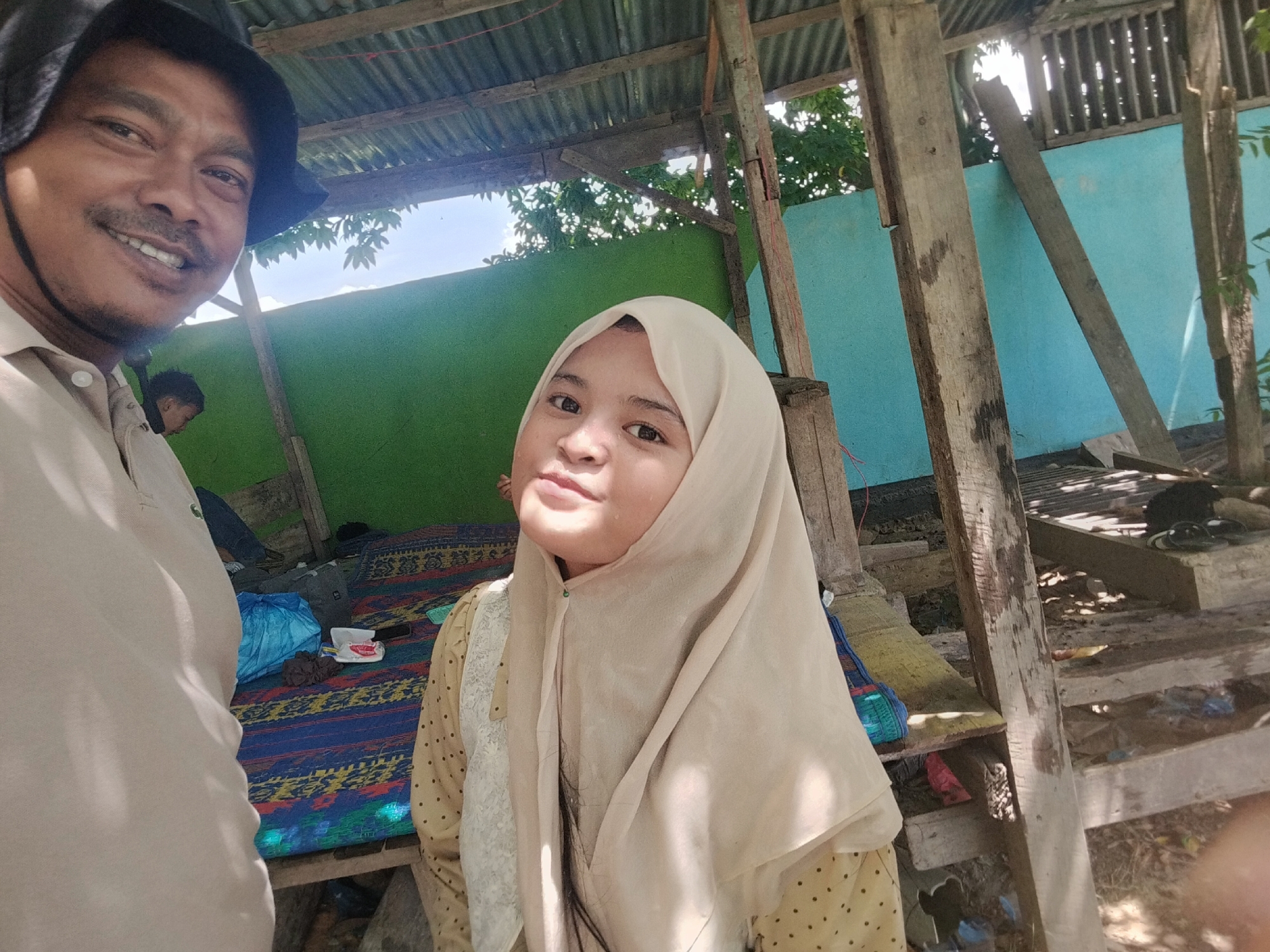 In Tegarmiko, Indonesia, a man wearing a dark bucket hat and a light-colored polo shirt is visible on the left side of the image. He is smiling and looking towards the camera. To his right, a young woman wearing a beige hijab and a patterned yellow and black dress is also looking towards the camera. The background shows a simple structure with a corrugated metal roof supported by wooden beams. A green wall is behind the people, and to the right, a bright blue wall is visible. The ground is covered with a mat featuring a colorful, intricate pattern. Various items, including bags and what appear to be personal belongings, are scattered on the mat and on wooden shelves below. The overall impression is of an outdoor, informal setting, possibly a home or a communal area in a tropical environment.