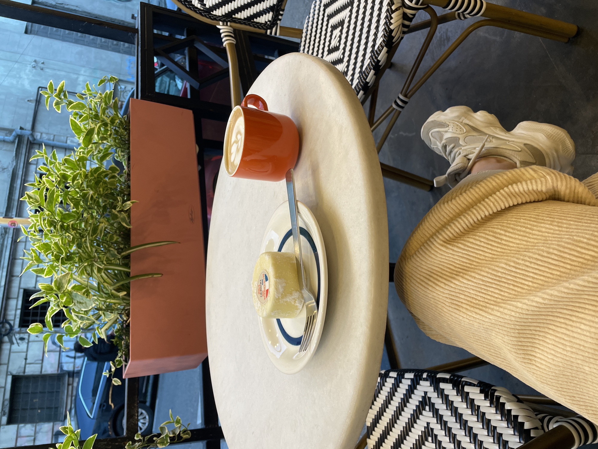A high-angle, slightly rotated shot captures a person enjoying coffee and dessert at a cafe table in Tbilisi, Georgia. The main subjects are an orange coffee mug with delicate latte art, a small cream roll or dessert on a white plate with blue trim, and a silver fork. These items rest on a light-colored, possibly marble, oval table.
Partially visible is the person's lower leg, clad in light beige corduroy pants, and a foot wearing a chunky white and light grey sneaker. The seating consists of chairs with distinctive black and white woven rattan-style backs and seats.
To the left of the table, a rectangular, terracotta-colored planter holds a lush green plant with variegated leaves. Beyond the plant, a window or reflective surface offers a glimpse of an urban street scene, showing reflections of buildings with windows and a dark blue car, suggesting the cafe is situated street-side. The bright lighting indicates it is daytime.
On the dessert's wrapper or label, visible text includes "DESSERT," "ROULÉ," and "LÉGÈRE," accompanied by a small red, white, and blue logo that resembles a flag. The image conveys a casual moment of enjoyment at an urban cafe.