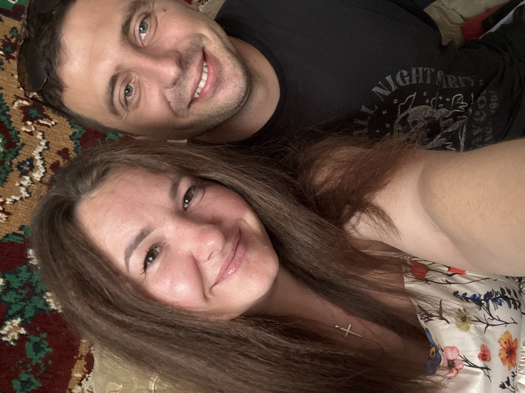 The image is a selfie of a man and a woman, taken indoors. The man is on the right, wearing a black t-shirt and sunglasses on his head. The woman is on the left, with long brown hair, wearing a floral dress and a cross necklace. They are both smiling. The background appears to be a tapestry or rug with a red, green, and brown patterned design. The lighting suggests it is daytime, possibly near a window. The location of Хаціслаўскі сельскі Савет, Belarus is not discernible from the image itself.