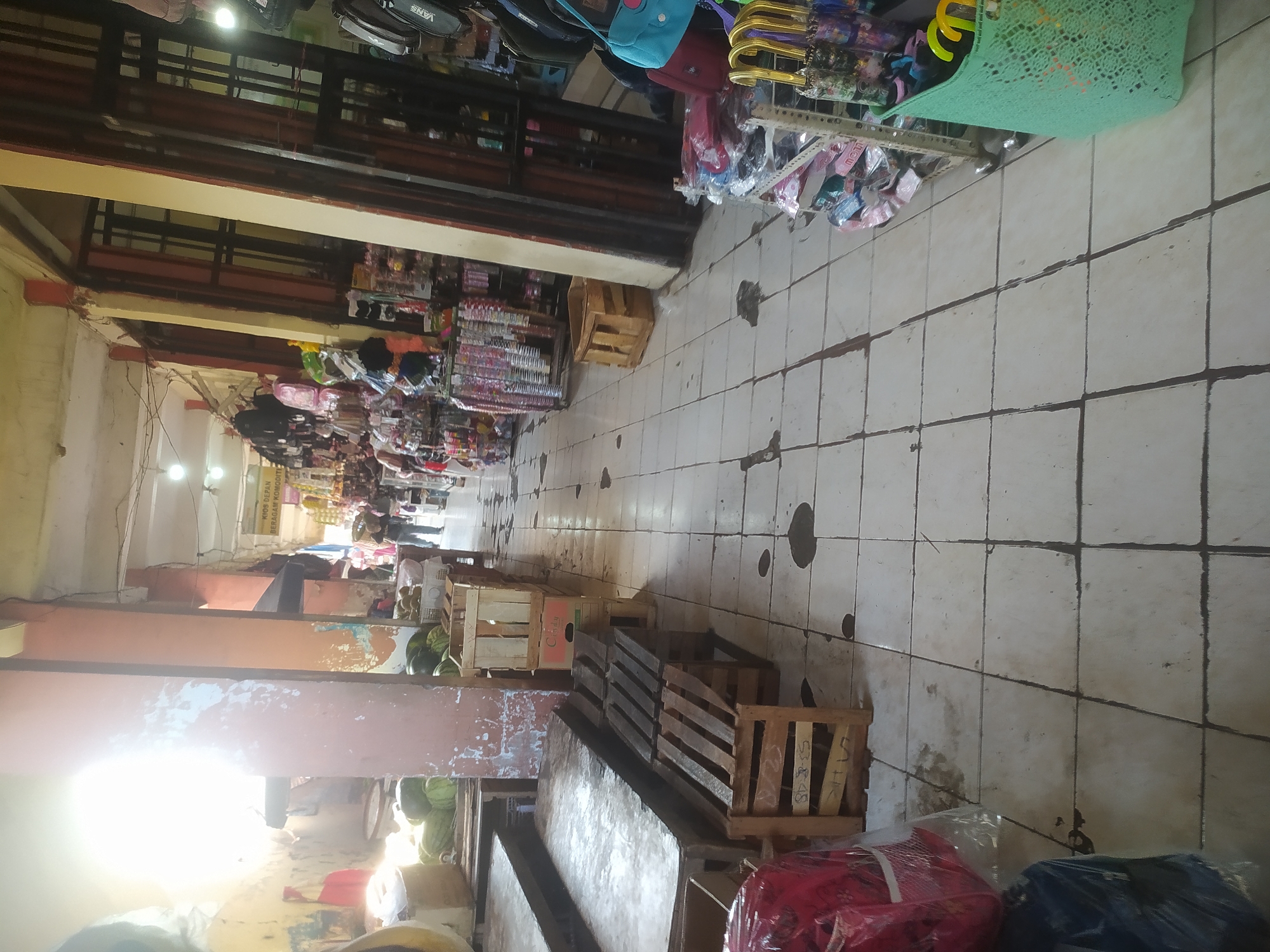 The image depicts a market scene in Kupang, Indonesia. The market appears to be an indoor or semi-covered structure with a tiled floor. Stalls are set up on both sides of a walkway, displaying various goods. On the left, there are fruits, possibly watermelons and durians, arranged in wooden crates. Further down the walkway, there are more crates and what appear to be benches or seating made of wooden slats.
On the right side of the walkway, the stalls are filled with consumer goods. These include clothing, bags, umbrellas, and other packaged items. Some stalls have racks displaying merchandise. The architecture suggests a functional, possibly older, market building with exposed beams and ceiling lights. Sunlight streams in from an opening on the left, creating a bright, washed-out area and casting shadows. A few individuals are visible walking through the market in the distance, suggesting it is open for business. The overall impression is of a bustling marketplace where goods are sold and people are going about their shopping.