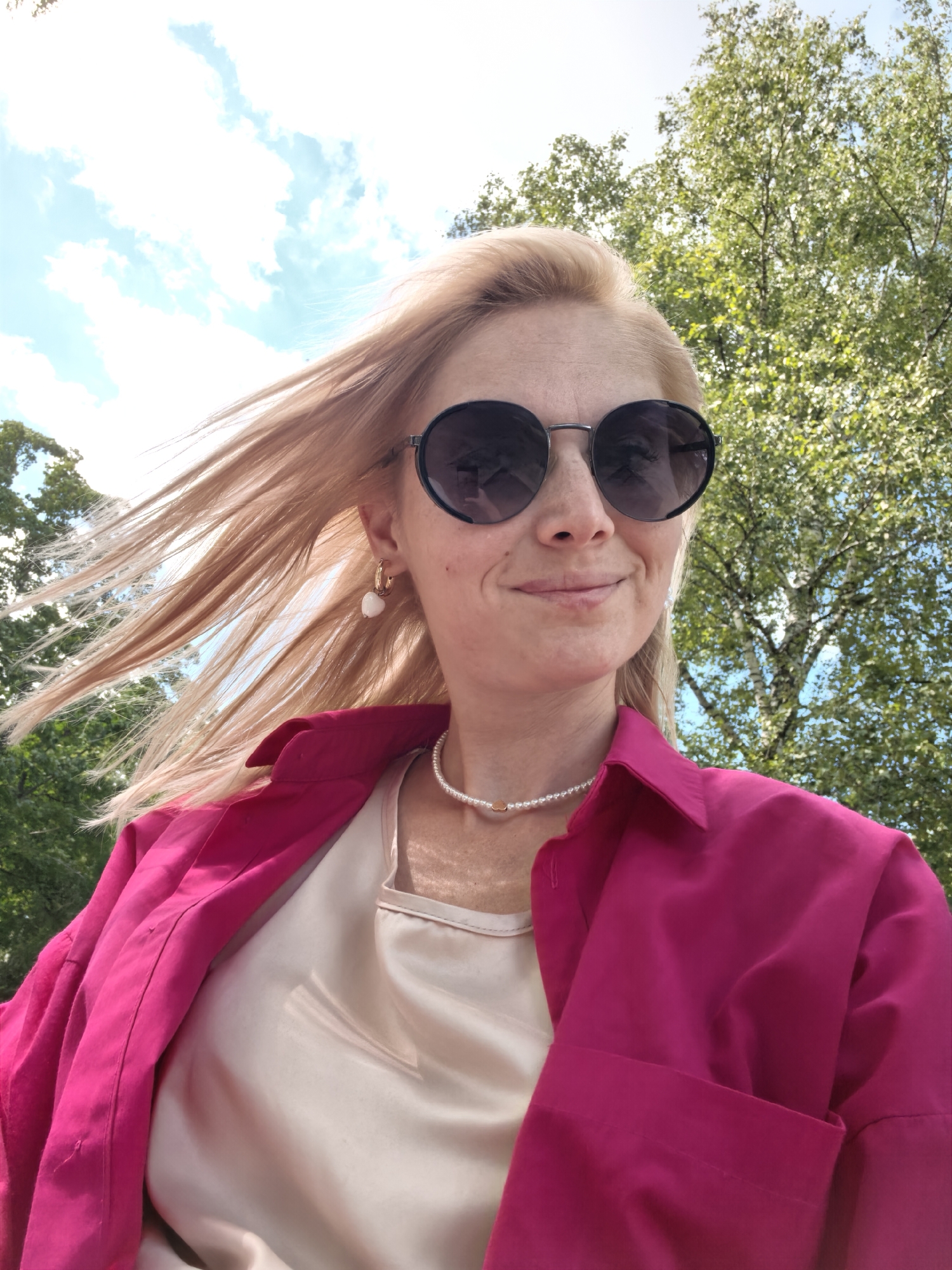 The image shows a woman with blonde hair, wearing sunglasses, a pink open shirt, and a light-colored camisole. She has a pearl necklace and heart-shaped earrings. The background consists of a bright blue sky with clouds and green trees. The image was taken outdoors on a sunny day. There is no information about the location in Sterlitamak, Russia.