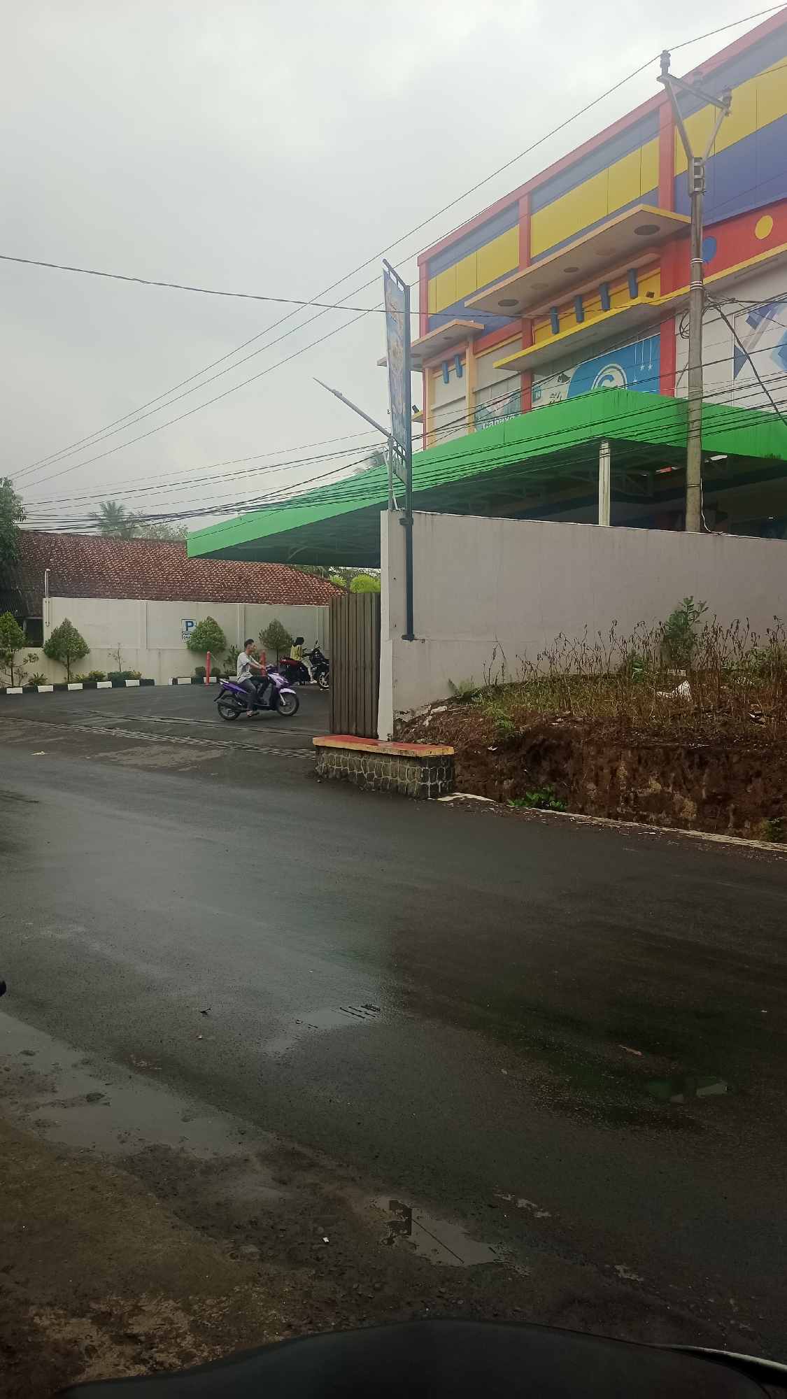 A street scene in Cikalong, Indonesia, shows a wet asphalt road in the foreground, reflecting the overcast sky. To the left, a paved area with white borders and small trees leads to a building with a red-tiled roof. Two motorcycles are parked near a wall, one with a rider wearing a purple shirt. Further back, a multi-story building with colorful facade elements in red, yellow, and blue is visible, featuring a prominent green awning. Power lines crisscross the sky above. The area appears to be a commercial or public space, possibly a parking lot or the entrance to a facility.