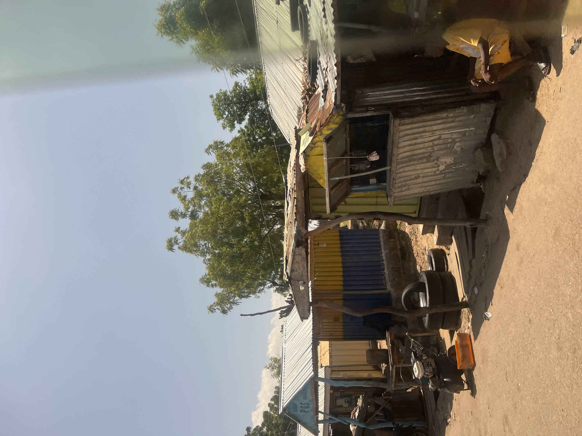 The image displays an informal commercial stretch within Kwara State, Nigeria, under a bright, clear sky. A row of four distinct, single-story structures, primarily constructed from corrugated metal sheets, lines an unpaved, light brown dirt road.
On the left, a stall with a reflective, corrugated metal roof features blue-painted wooden supports and a prominent sign reading "DIBABA POS AVAILABLE," indicating a Point of Sale service. Adjacent to this, a structure exhibits horizontal bands of bright yellow and dark blue corrugated metal. Further to the right, a smaller, yellow-fronted stall presents an open service window. Its corrugated metal roof is notably rusted and holds scattered organic debris, with a single bare light bulb suspended from its underside. The structure on the far right is composed of dark, weathered corrugated metal and is mostly obscured by shadow.
In the foreground and mid-ground, the unpaved path shows an orange plastic crate, and to its right, a stack of three black tires. A small, portable engine or generator on wheels is situated near the left-center.
On the far right, an individual with dark skin is seated in a low squat or on a low step next to the darkest stall. They are dressed in a bright yellow short-sleeved t-shirt and matching yellow shorts, paired with black slide sandals, and are looking towards the left.
The background features a clear, pale blue sky with sparse white clouds, above a dense line of green-leafed trees. Strong, defined shadows are cast on the ground and structures, consistent with direct overhead sunlight. A faint vertical light anomaly is present on the far right edge of the image.