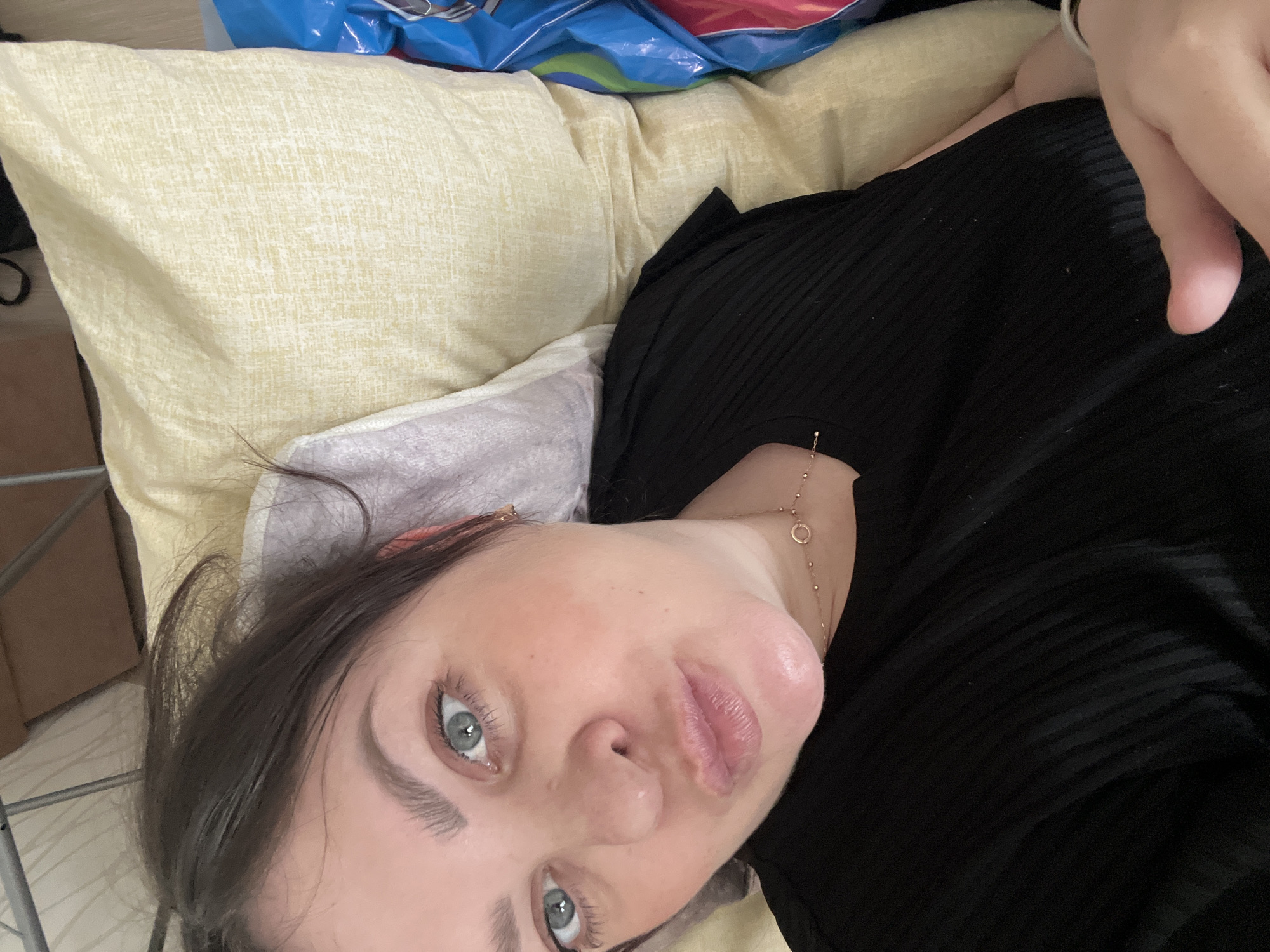 A woman with fair skin, brown hair, and blue eyes lies on a pillow. She wears a black shirt and a gold necklace. The pillow is yellow and textured. A blue and red plastic bag is visible in the background. The location is not discernible from the image.