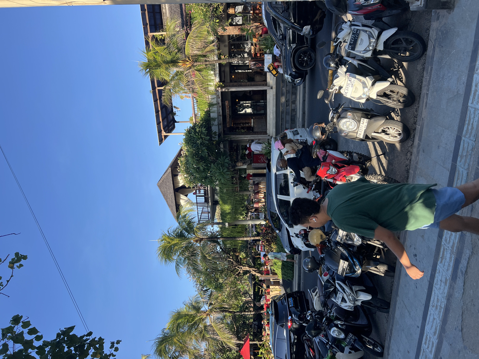 In Kuta, Indonesia, a street scene unfolds under a clear blue sky. Several parked motorbikes line the foreground, with a man in a green t-shirt and blue shorts walking away from the camera. Behind the motorbikes, a row of cars, including black SUVs and a white van, are parked. Further back, a two-story building with an open-air upper level, lined with tables and chairs, suggests a restaurant or cafe. Palm trees and other lush greenery surround the building and extend along the street. People can be seen in the background, some standing near the building and others seated at tables. The scene captures a typical day in a busy urban area with a tropical atmosphere.
