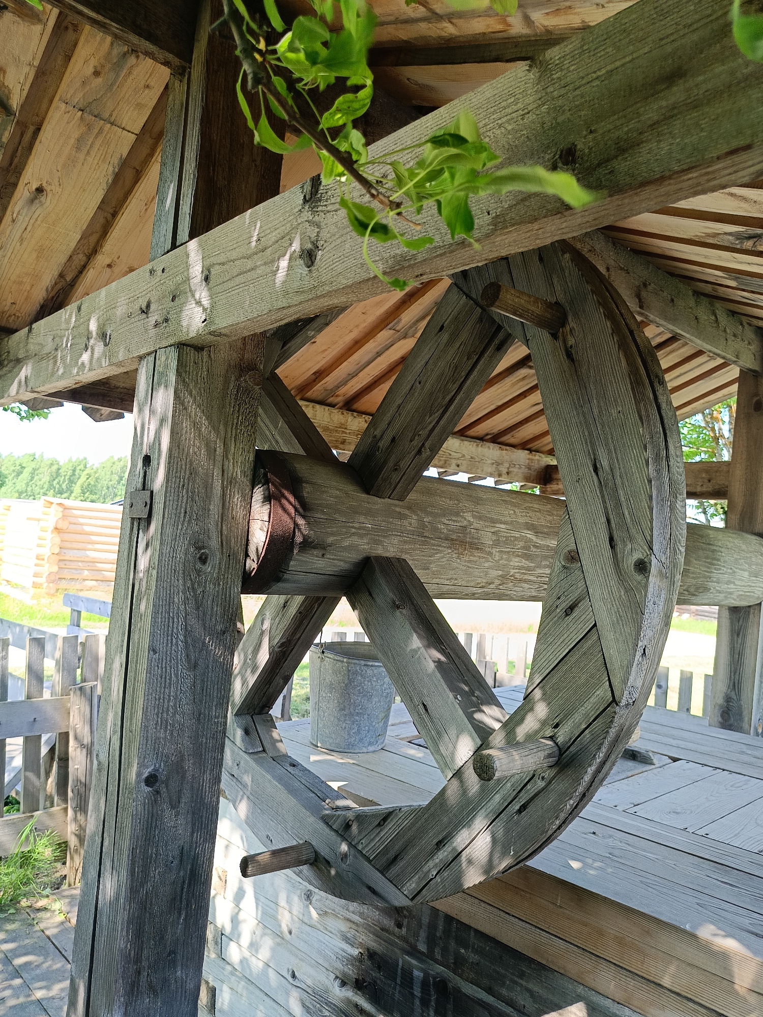 The image shows a close-up view of an old wooden well with a large wooden wheel used for drawing water. The well structure is made of weathered, grey wood, with visible grain and knot patterns. The wheel has spokes and a rim, and it's supported by a thick wooden axle. A metal bucket is visible on the wooden platform surrounding the well. In the background, a wooden fence and a rustic log building are visible, suggesting a rural or historical setting. Green foliage hangs into the frame from above. The scene appears to be outdoors on a sunny day, with dappled sunlight casting shadows on the wooden surfaces. The overall impression is one of age and rustic charm. The location is identified as Popovka, Russia.