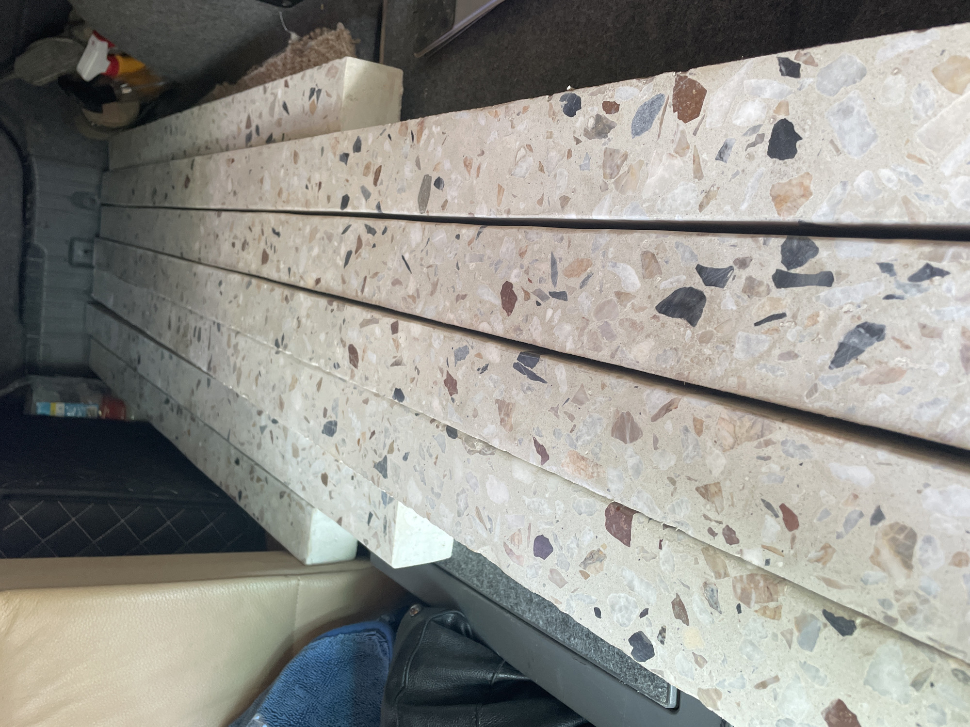 The image shows a stack of several light-colored terrazzo tiles with a speckled pattern of stones in various colors (brown, gray, white, black) resting on white blocks in what appears to be the cargo area of a vehicle. Parts of the vehicle's interior are visible, including gray carpeting/trim at the top of the frame, a light tan leather seat, and miscellaneous items. It is not possible to determine if this is a specific location in Krasnodar, Russia, from this image alone.